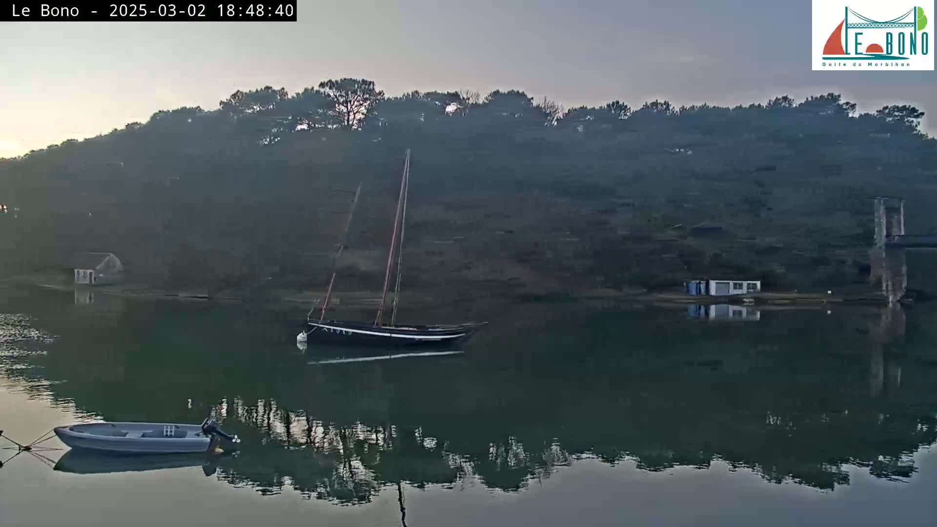 A sailboat and a small motorboat are moored in calm water next to a wooded shore under a dusky sky.