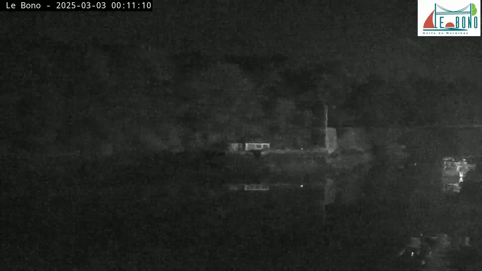 A low-light nighttime image shows a dark body of water reflecting a dark shoreline with some indistinct structures.