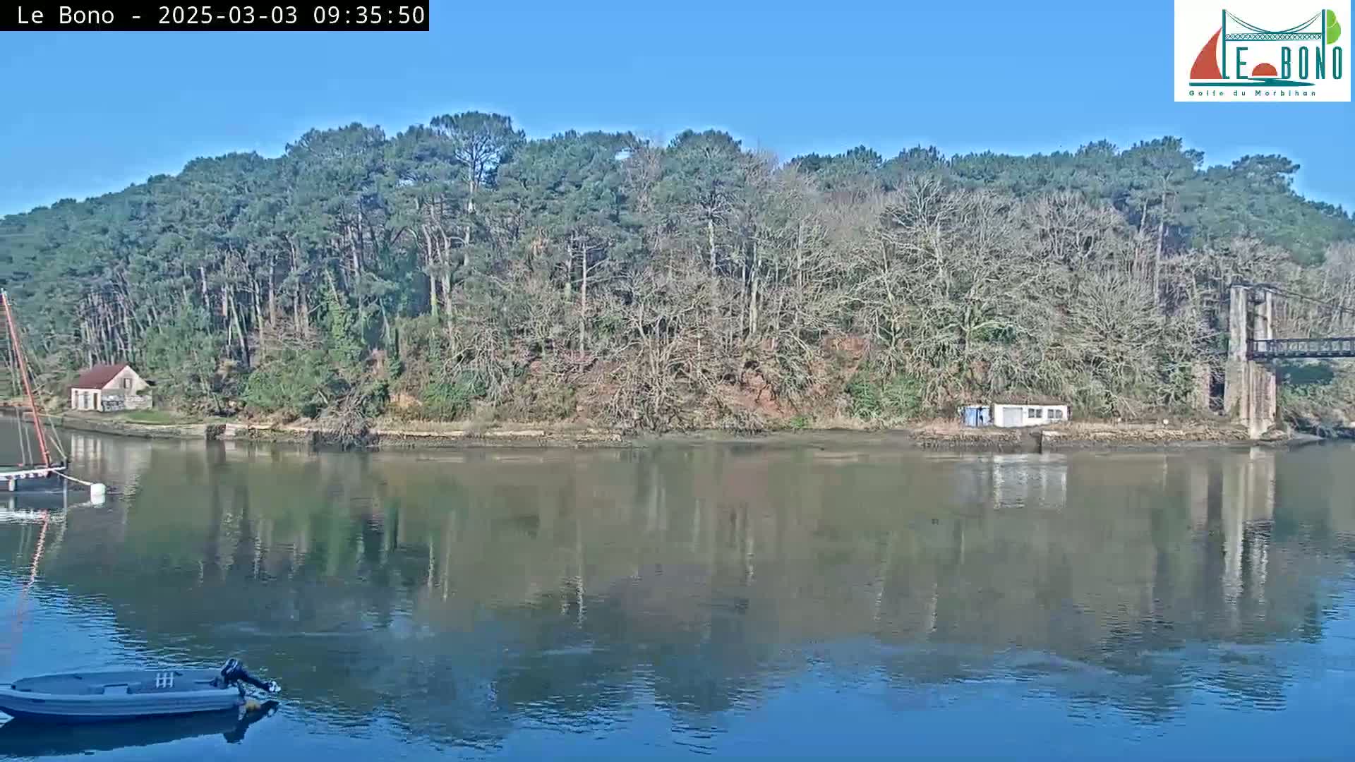 A calm river reflects a treeline and a suspension bridge under a clear blue sky.