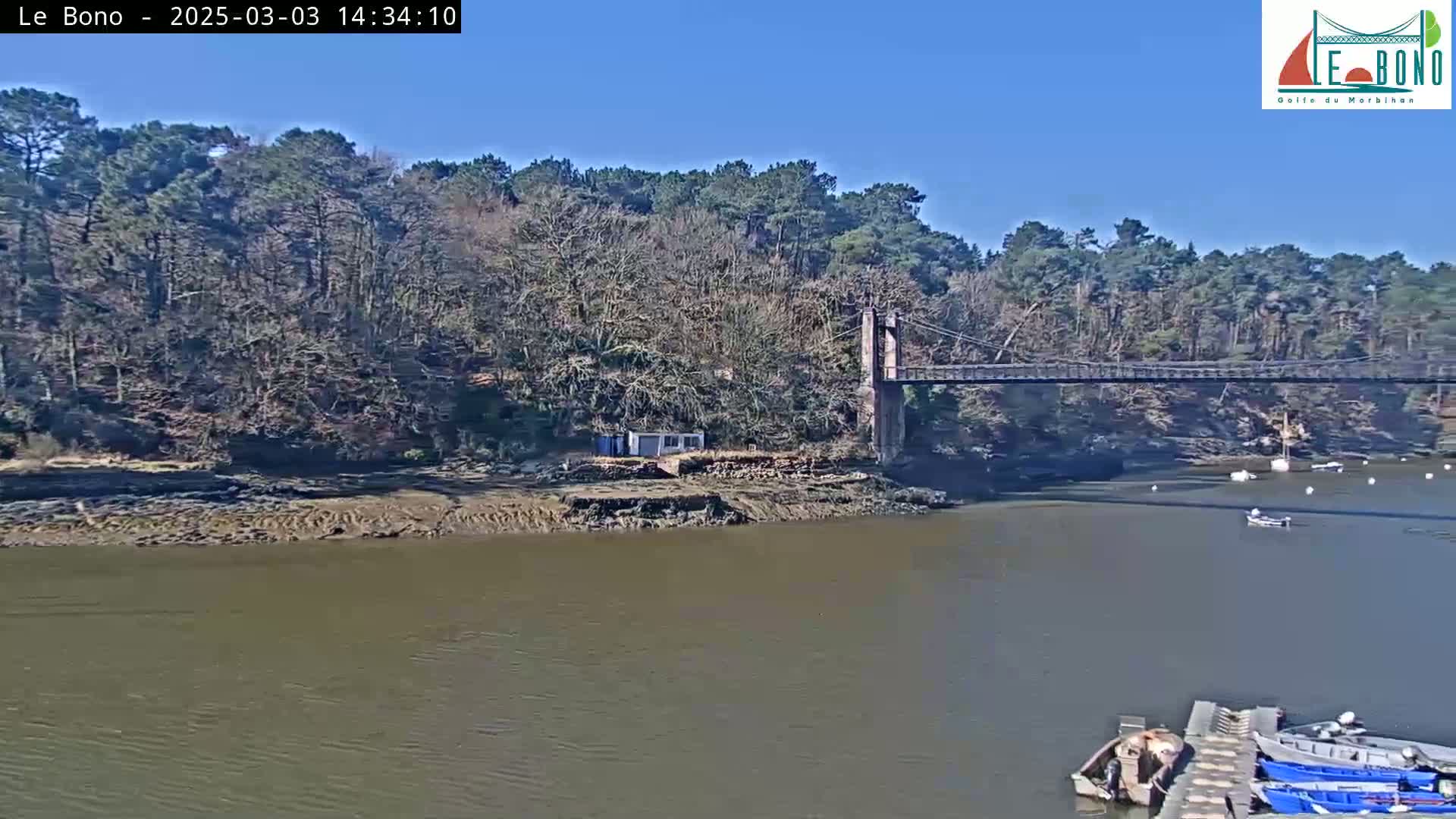 A suspension bridge spans a calm river under a clear blue sky, with bare trees lining the riverbanks and several small boats moored at a dock.