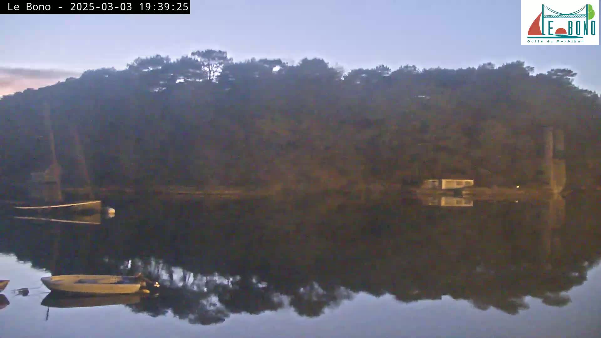 A calm body of water reflects a dark, tree-covered shoreline at dusk, with several small boats moored near the shore.