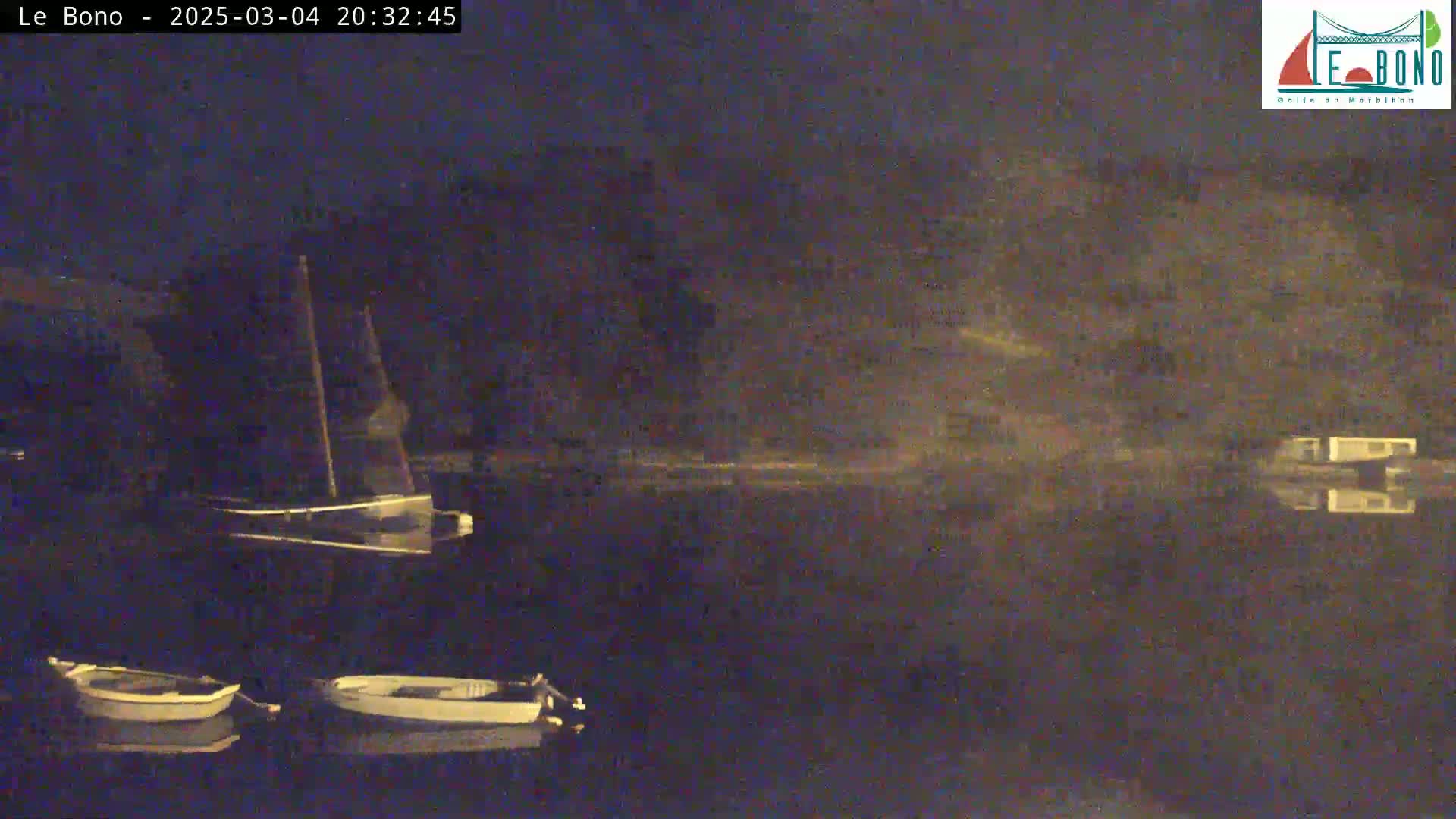 At night, three small boats are moored on a calm body of water, with a larger sailboat visible behind them.