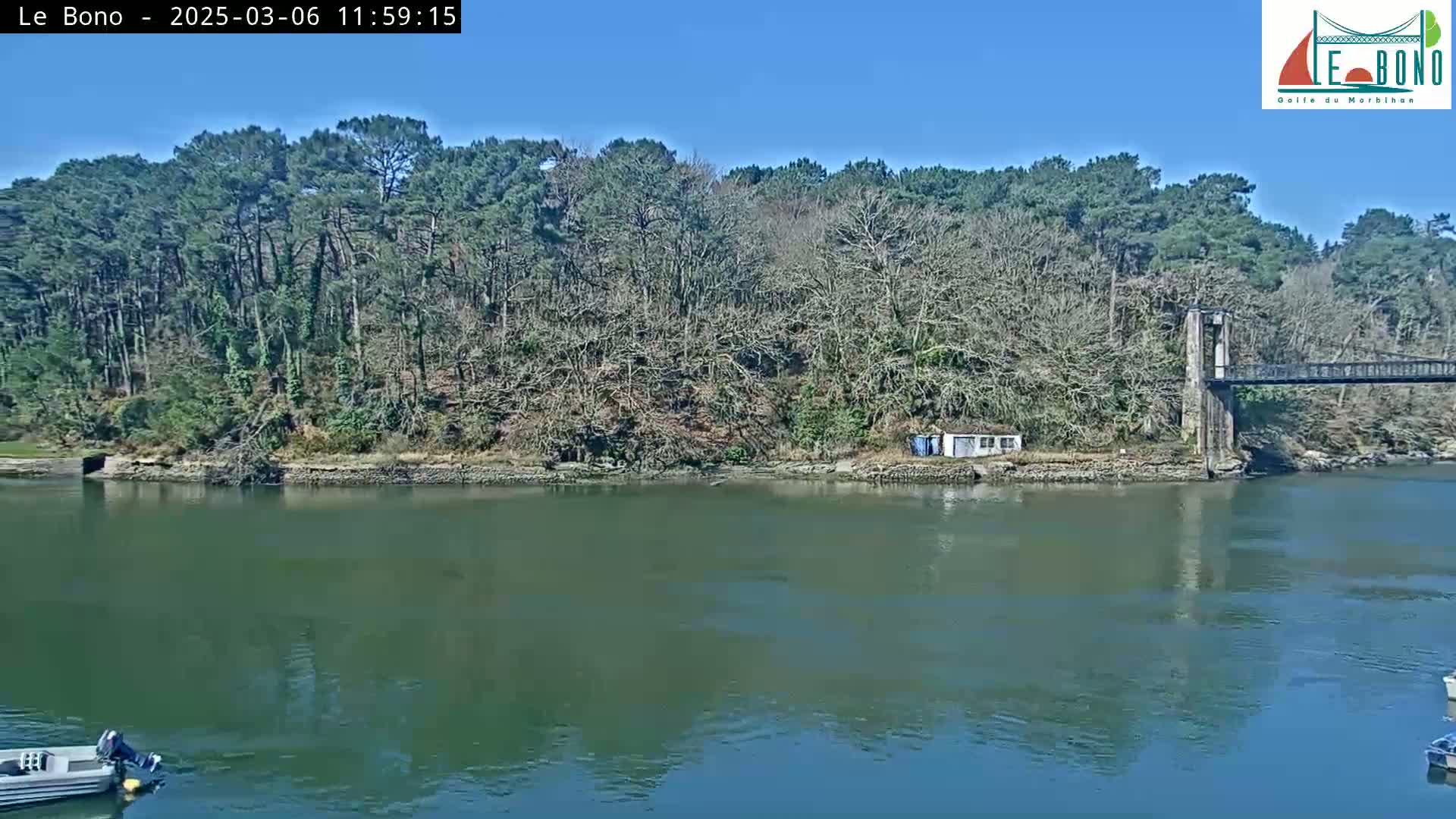 A calm river flows past a wooded shoreline with a small building and a suspension bridge in the background under a clear blue sky.