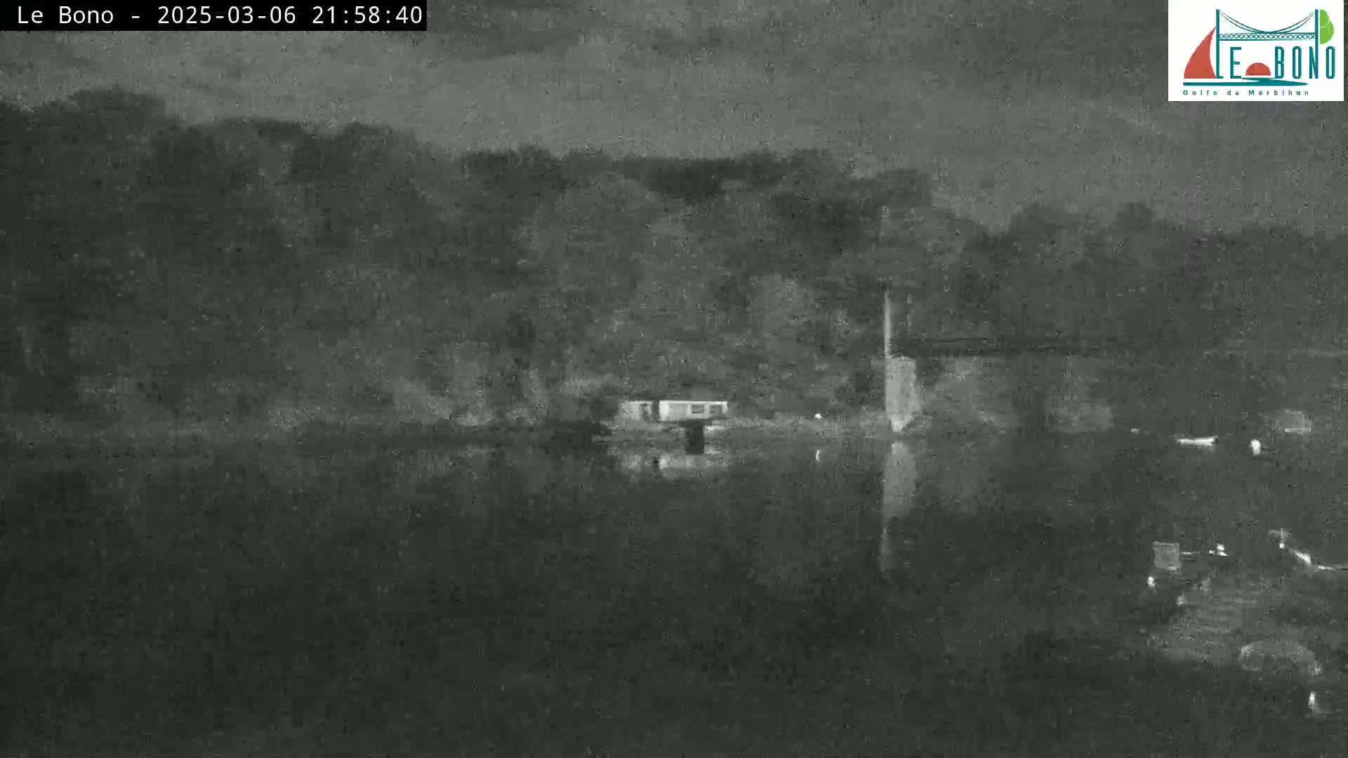 A dark, nighttime view of a calm body of water reflecting a treeline and a small structure on the far shore under an overcast sky.