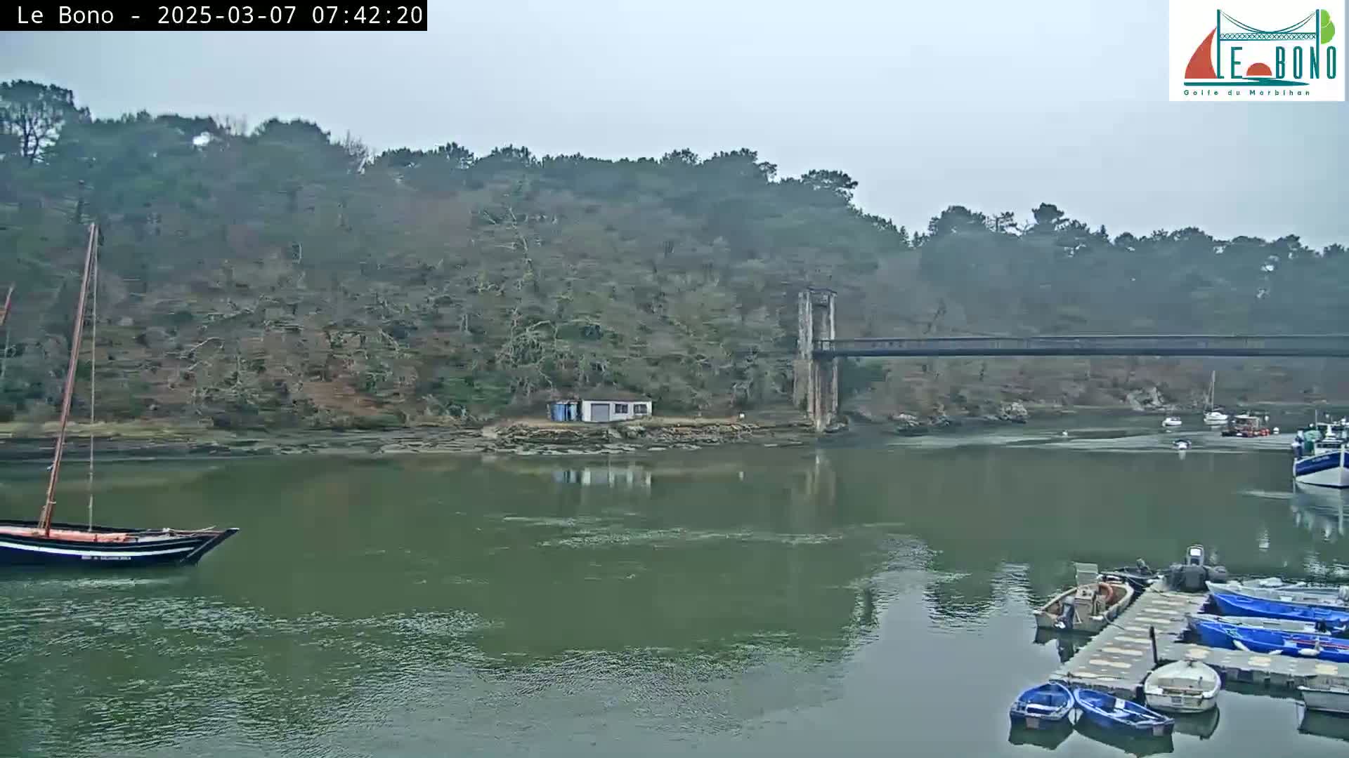 A calm, overcast day shows a river with a small sailboat and several small motorboats docked near a bridge spanning a narrow waterway, with a small building on the far shore and a lush green hillside behind it.