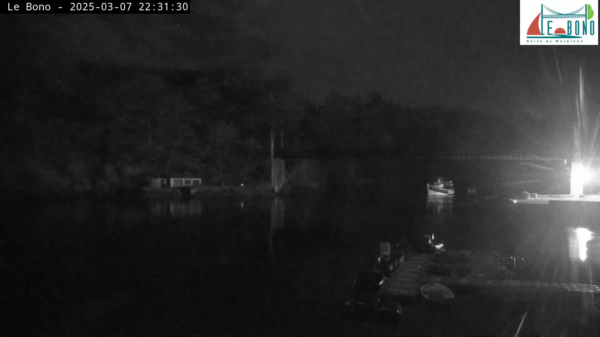 A nighttime, low-light view shows a dark body of water with a small boat and the dark silhouette of land and structures under a clear, dark sky.