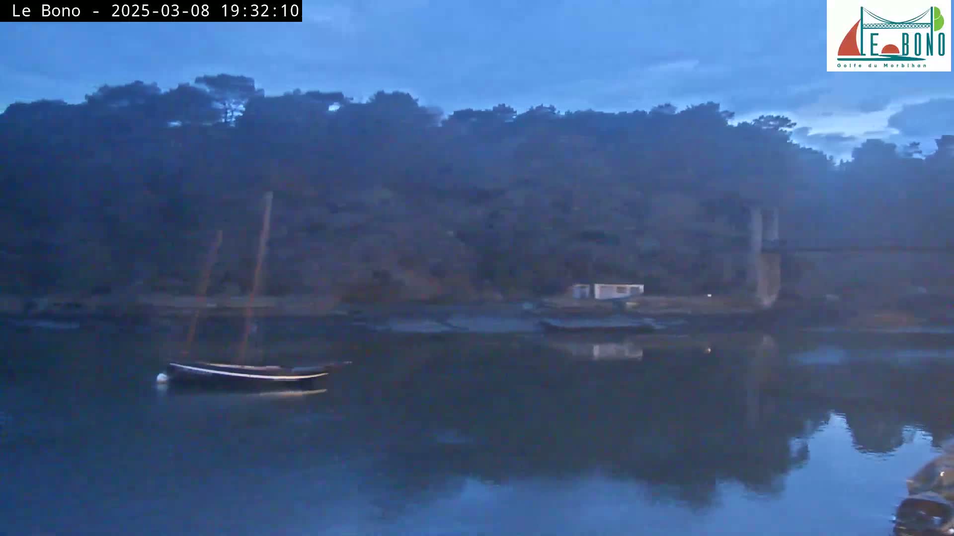 A small sailboat is moored in a calm, dark-blue body of water at dusk, with a line of trees and a structure on the far shore.