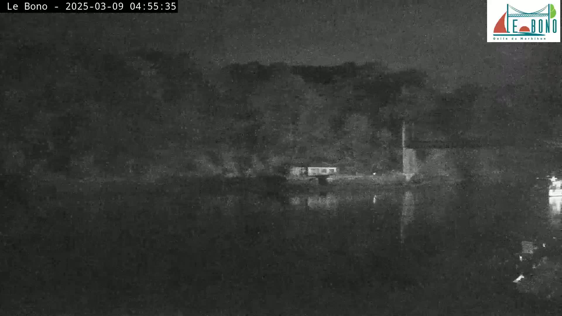 A nighttime, low-light image shows a dark body of water reflecting a dark shoreline with some structures and trees under a dark sky.
