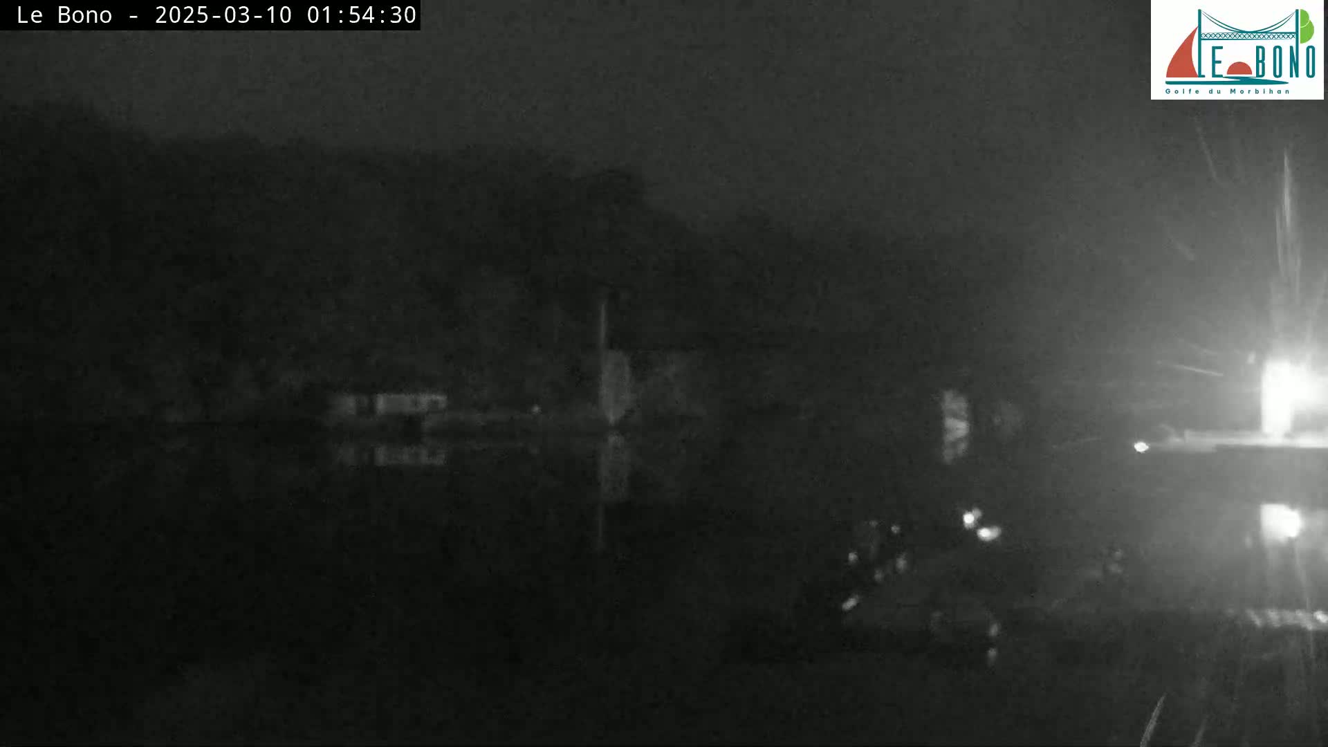 A nighttime, low-light image shows a dark body of water reflecting a dark shoreline under a seemingly rainy sky.