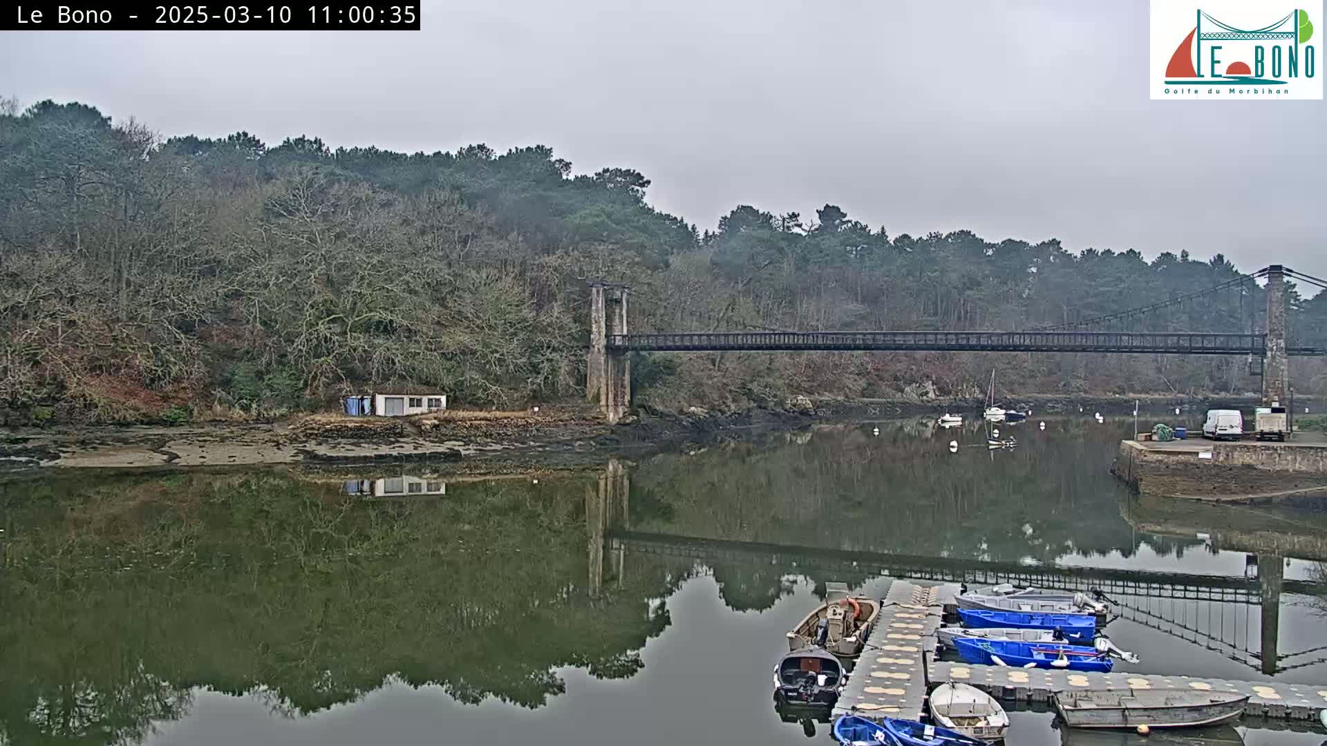 A suspension bridge spans a calm river reflecting the overcast sky, with several small boats moored at a dock in the foreground.