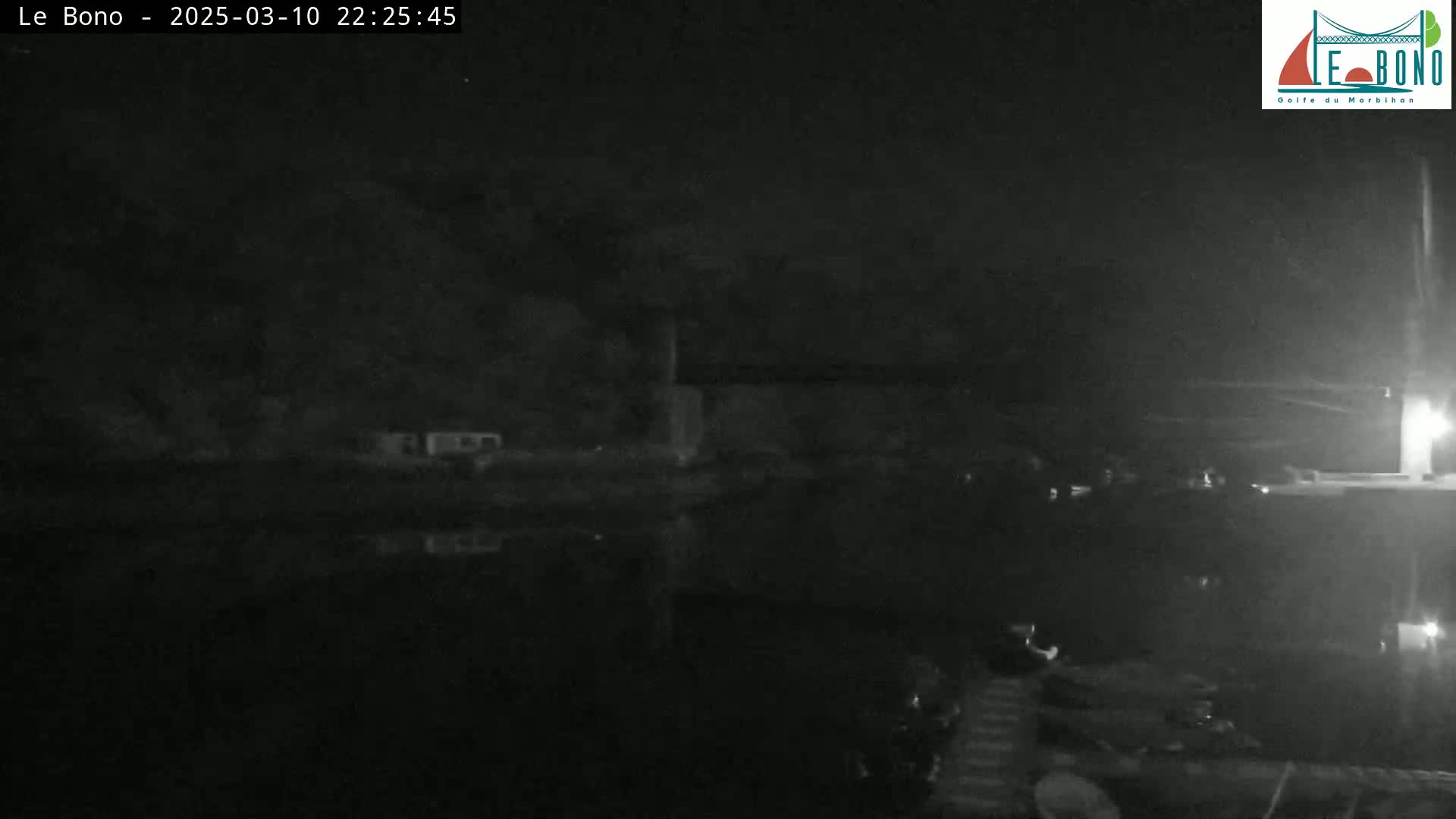 A nighttime, low-light image shows a dark body of water reflecting a dark shoreline with some structures and vehicles faintly visible.