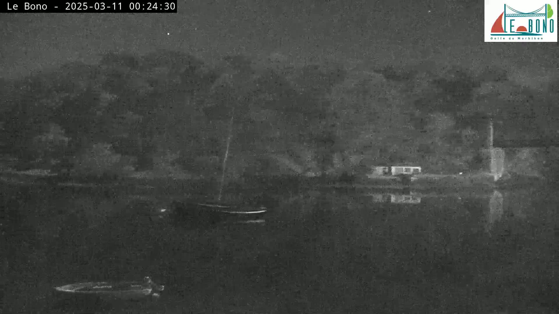 A nighttime, low-light image shows a calm body of water reflecting the dark shoreline where trees and buildings are visible; small boats are moored on the water.