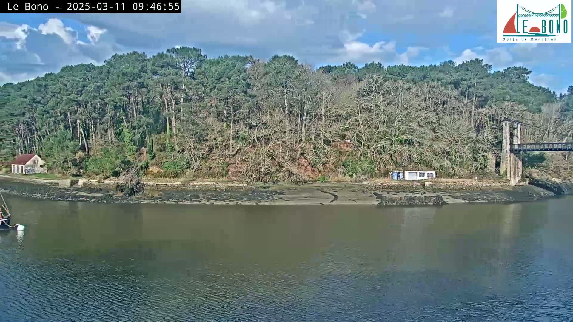 A mostly sunny day shows a small sailboat in a calm river, which runs alongside a wooded shoreline featuring a small building, and a suspension bridge in the background.