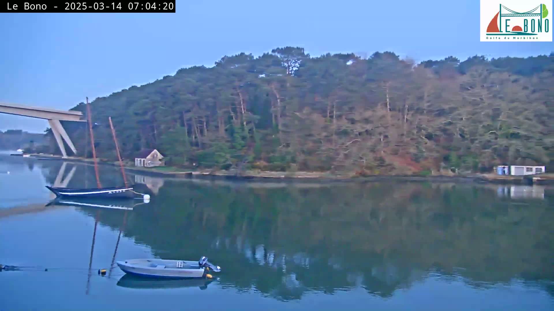 A calm body of water reflects a wooded shoreline and two small boats under a clear, pale sky.