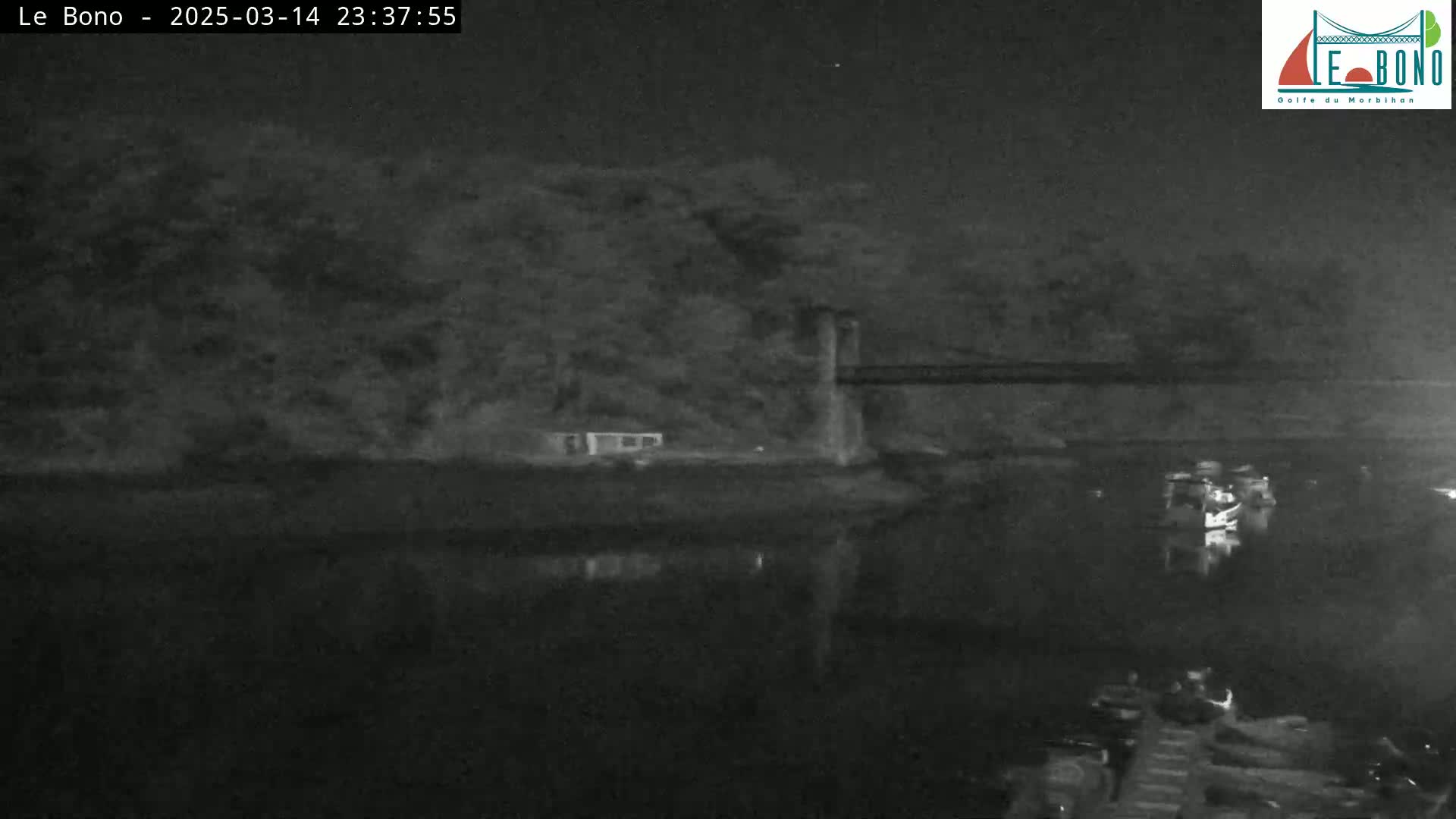 A nighttime, low-light image shows a dark body of water reflecting a suspension bridge and trees under a clear night sky.