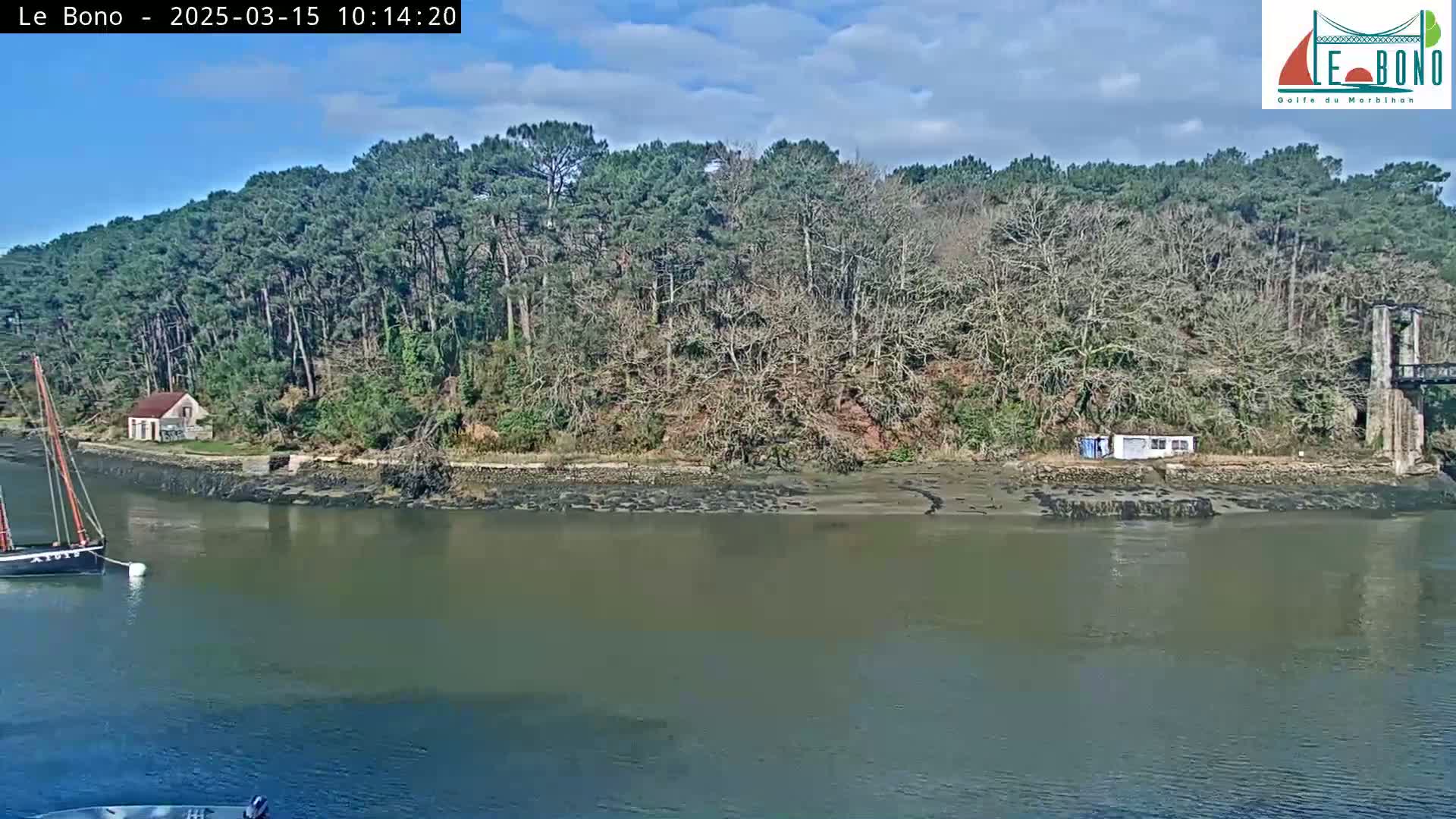 A small sailboat is moored in a calm river next to a wooded shoreline with a small building, under a partly sunny sky.