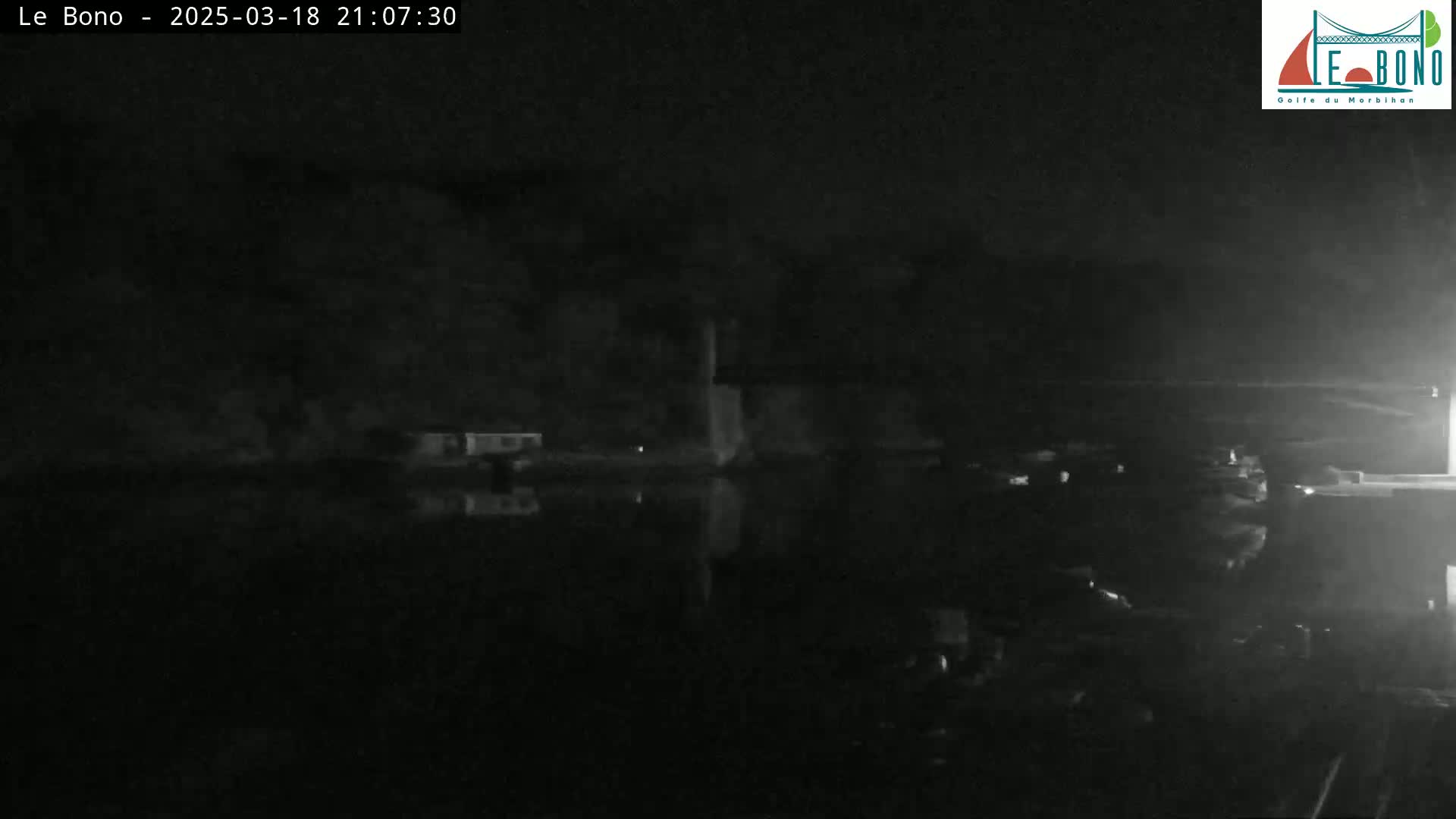 A nighttime, low-light image shows a dark body of water reflecting a dimly lit shoreline with some structures.