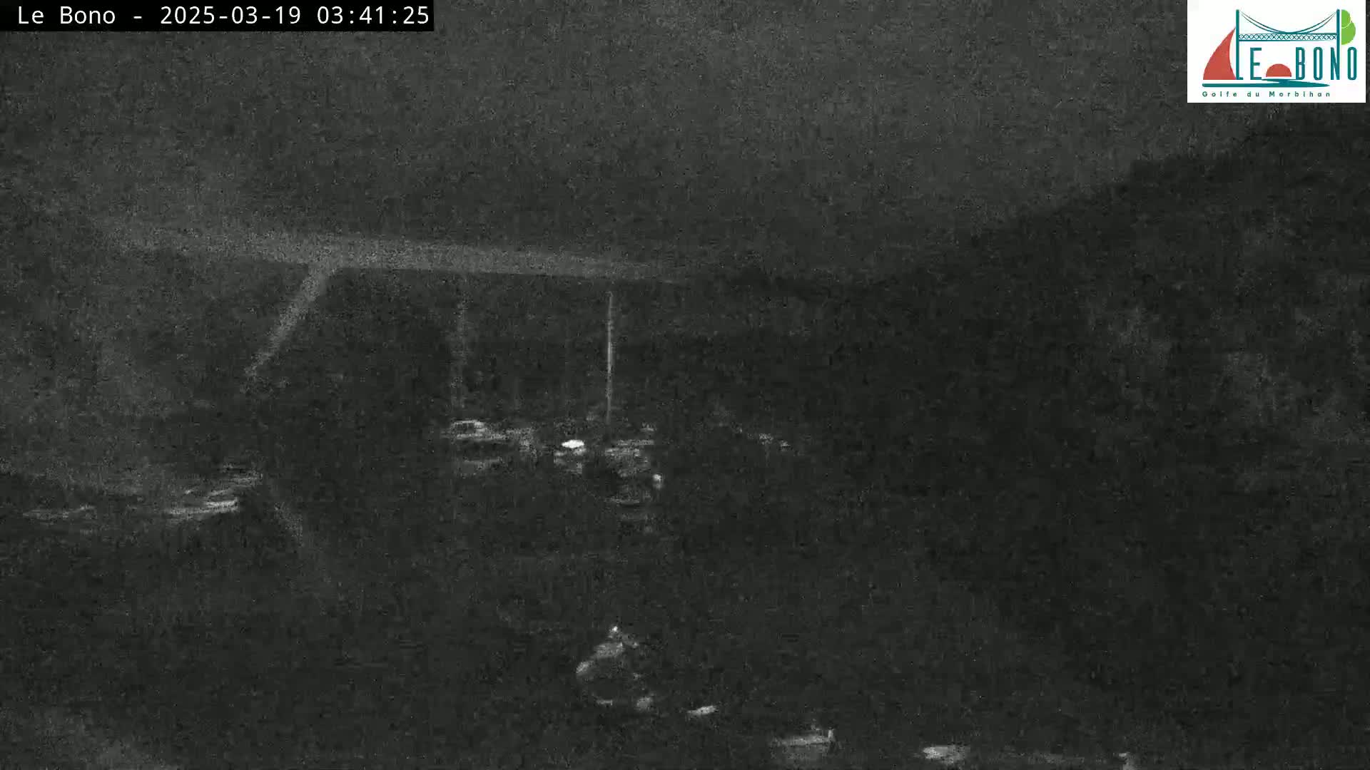 A nighttime, low-light image shows a dark, mostly empty area with a few indistinct objects and what appears to be a road or structure in the distance.