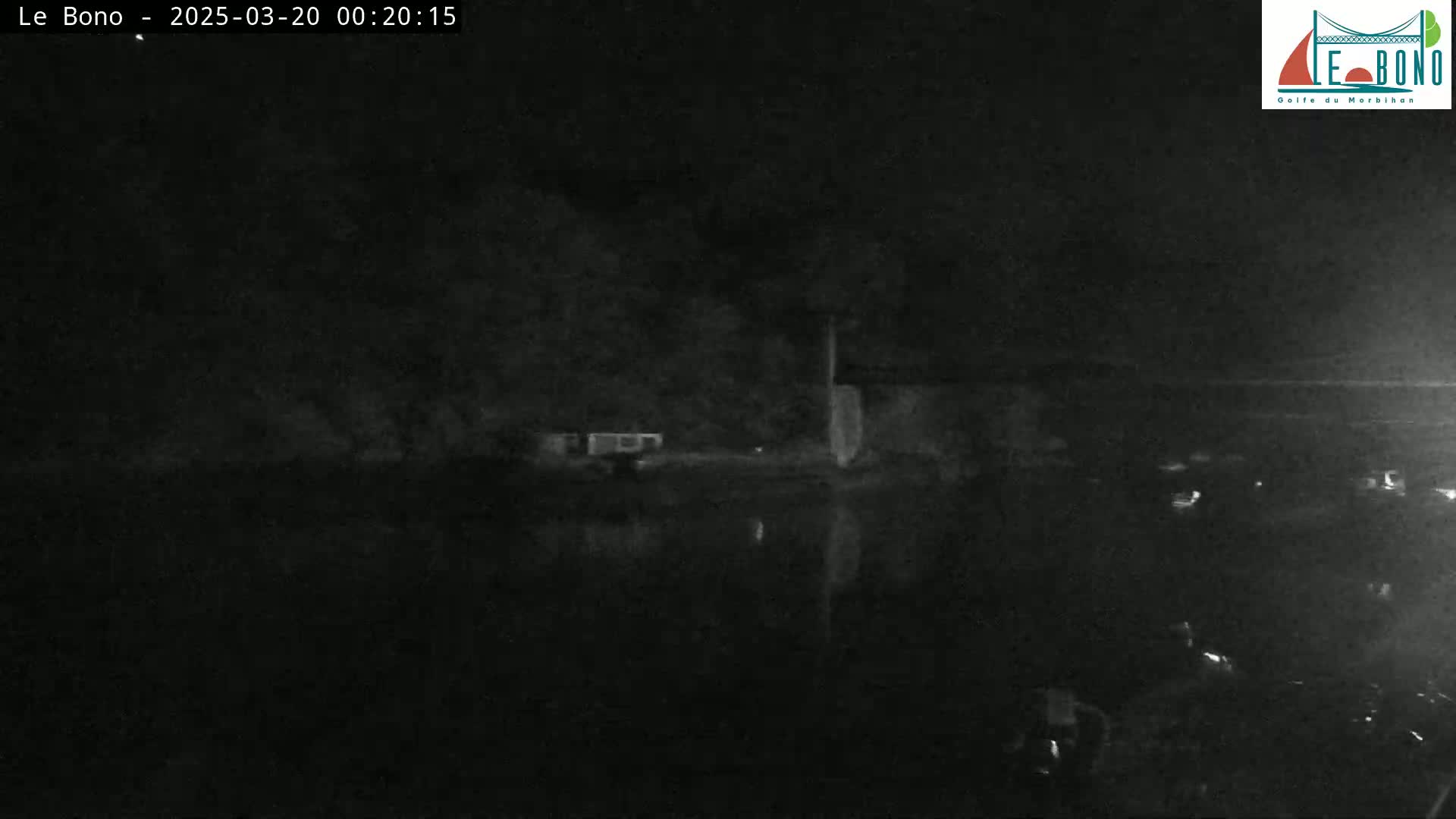 A nighttime, low-light image shows a dark body of water reflecting a dark shoreline with sparse structures under a clear, dark sky.