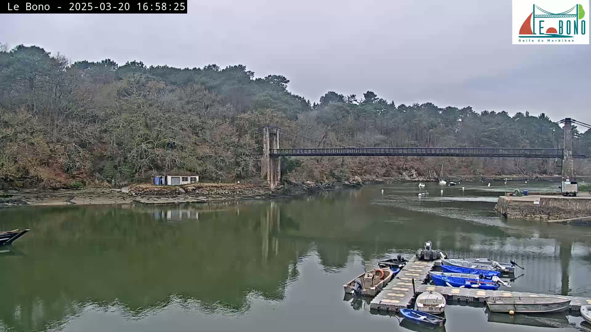 A suspension bridge spans a calm river, bordered by trees under an overcast sky, with several small boats moored at a dock.
