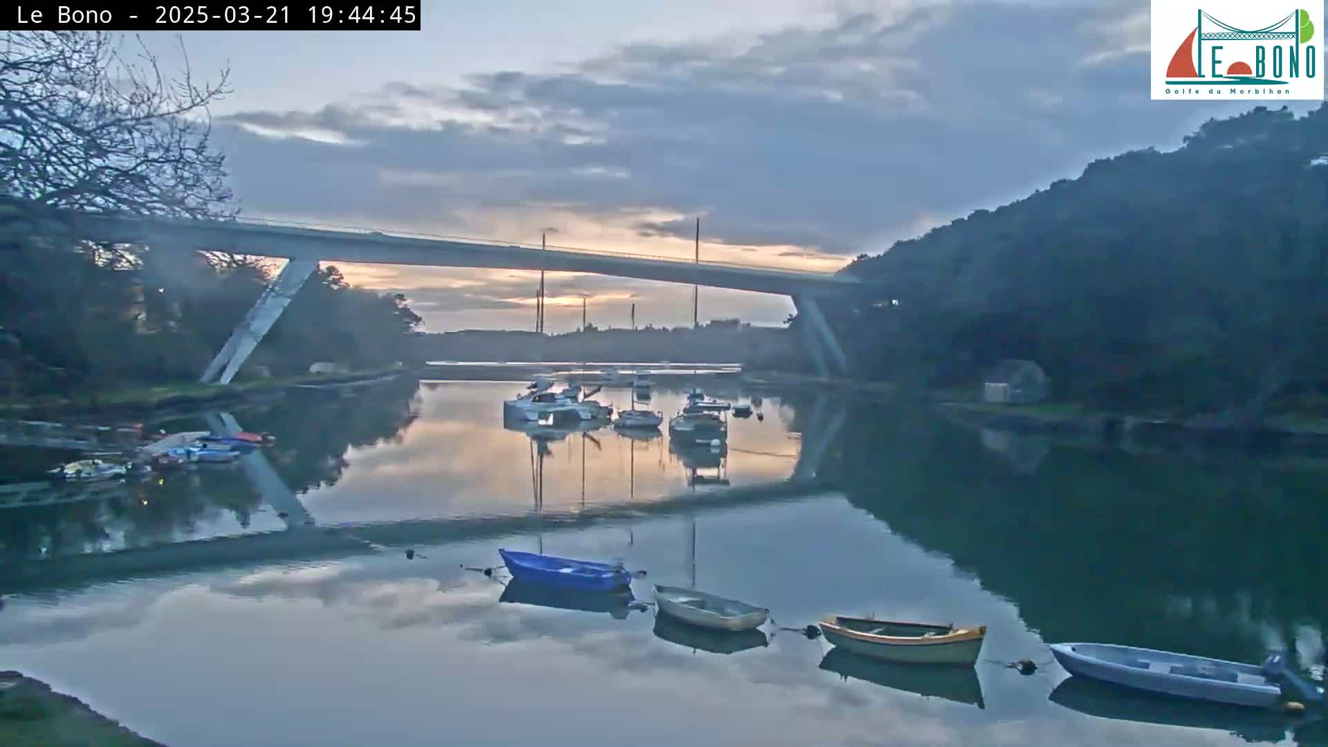 Several small boats are moored in a calm body of water under a modern bridge at dawn, with a cloudy sky above.