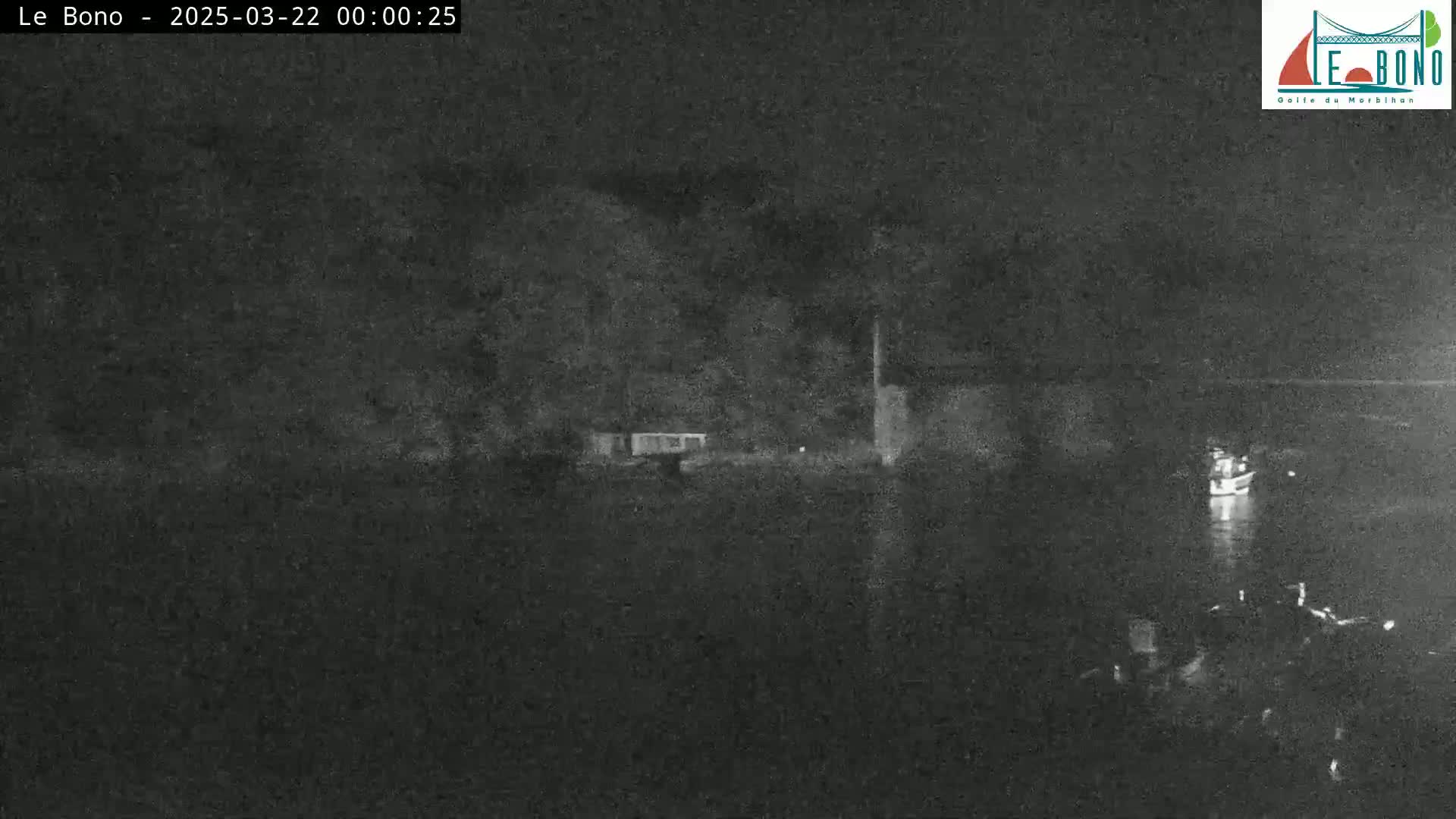 A nighttime, low-light image shows a dark body of water with a few small boats and a shoreline with some structures and trees.