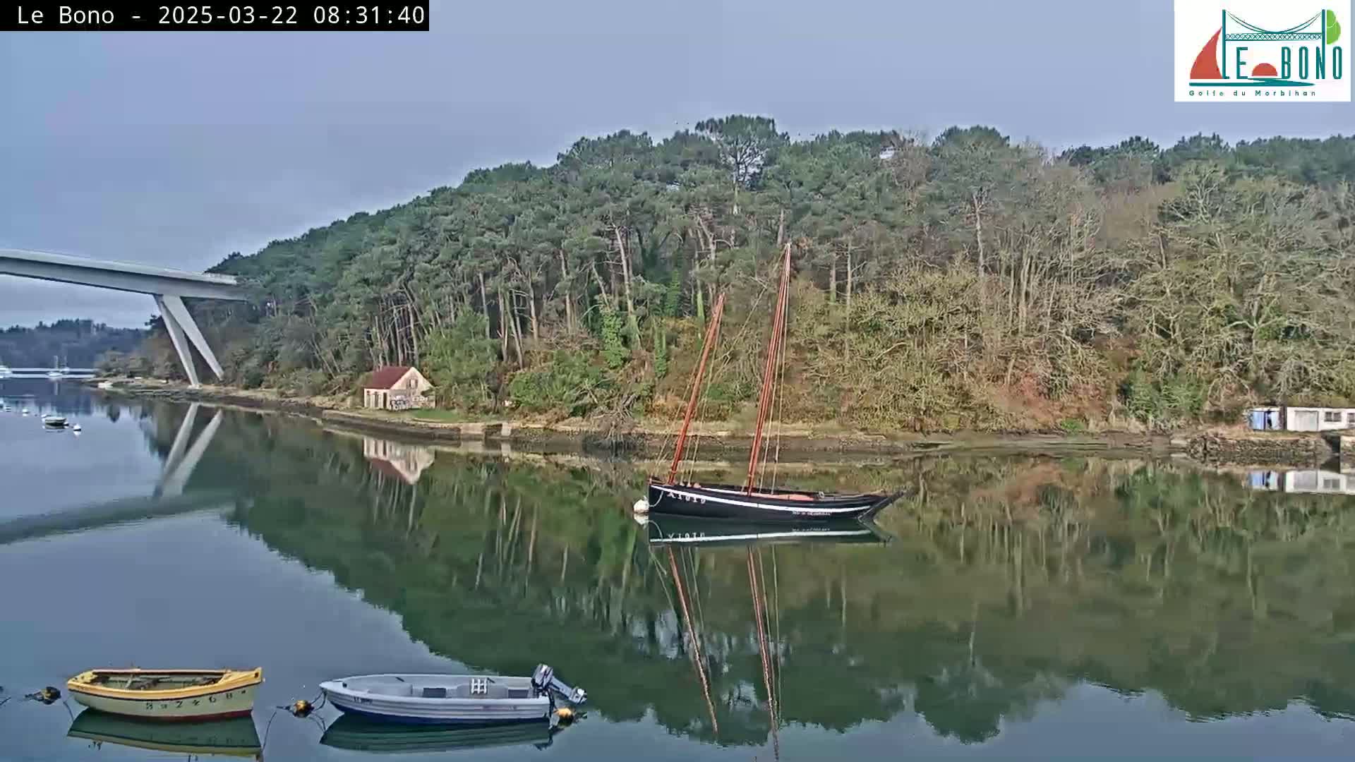 A calm body of water reflects a wooded shoreline, a sailboat, and a bridge, with two small motorboats in the foreground under a mostly clear sky.