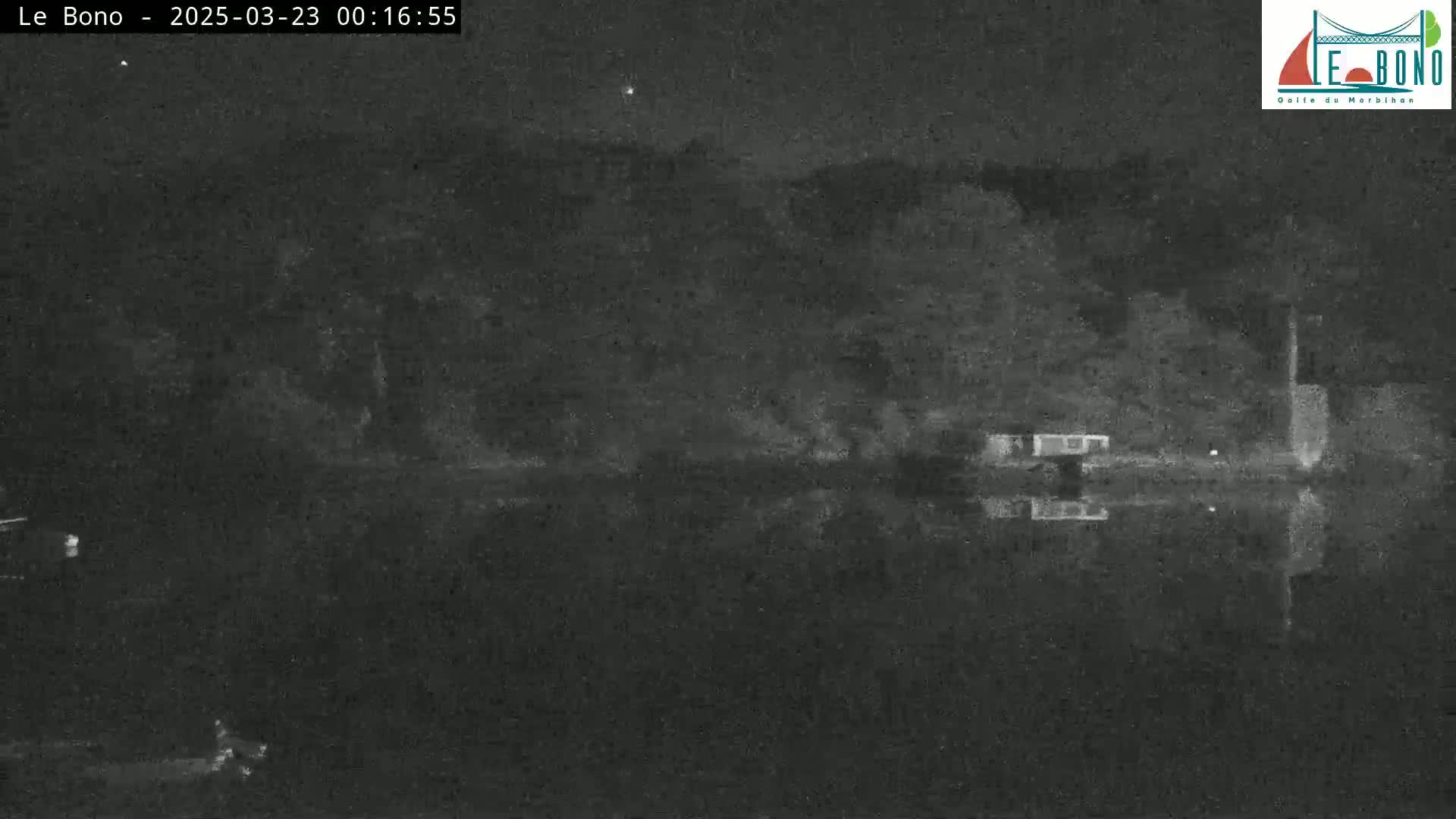 A nighttime, low-light image shows a dark body of water reflecting a dark shoreline with some indistinct structures under a clear, dark sky.