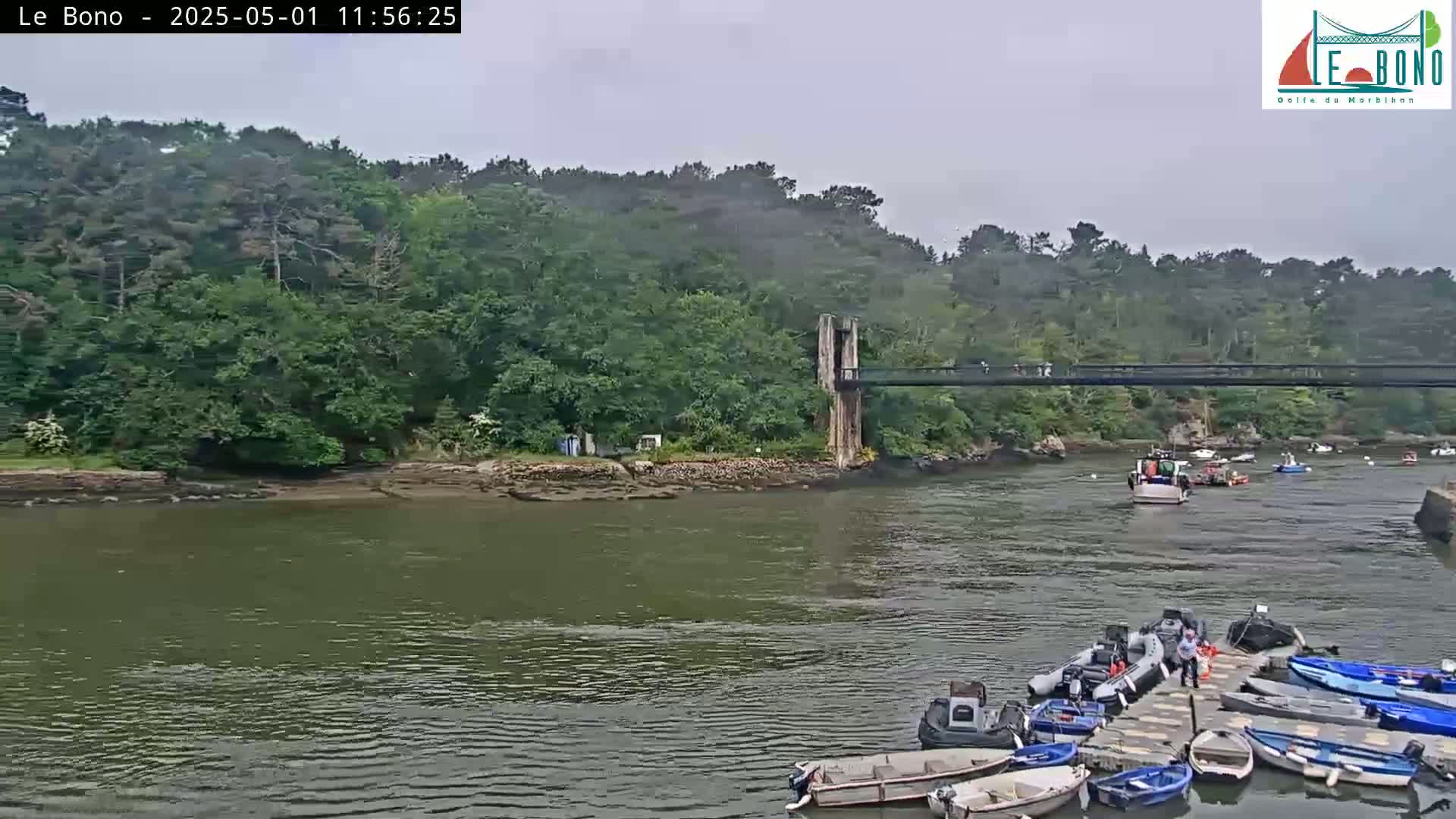 A calm river is shown with several small motorboats docked, a larger fishing boat in the river, and a pedestrian bridge spanning a narrow waterway lined with lush green trees under an overcast sky.