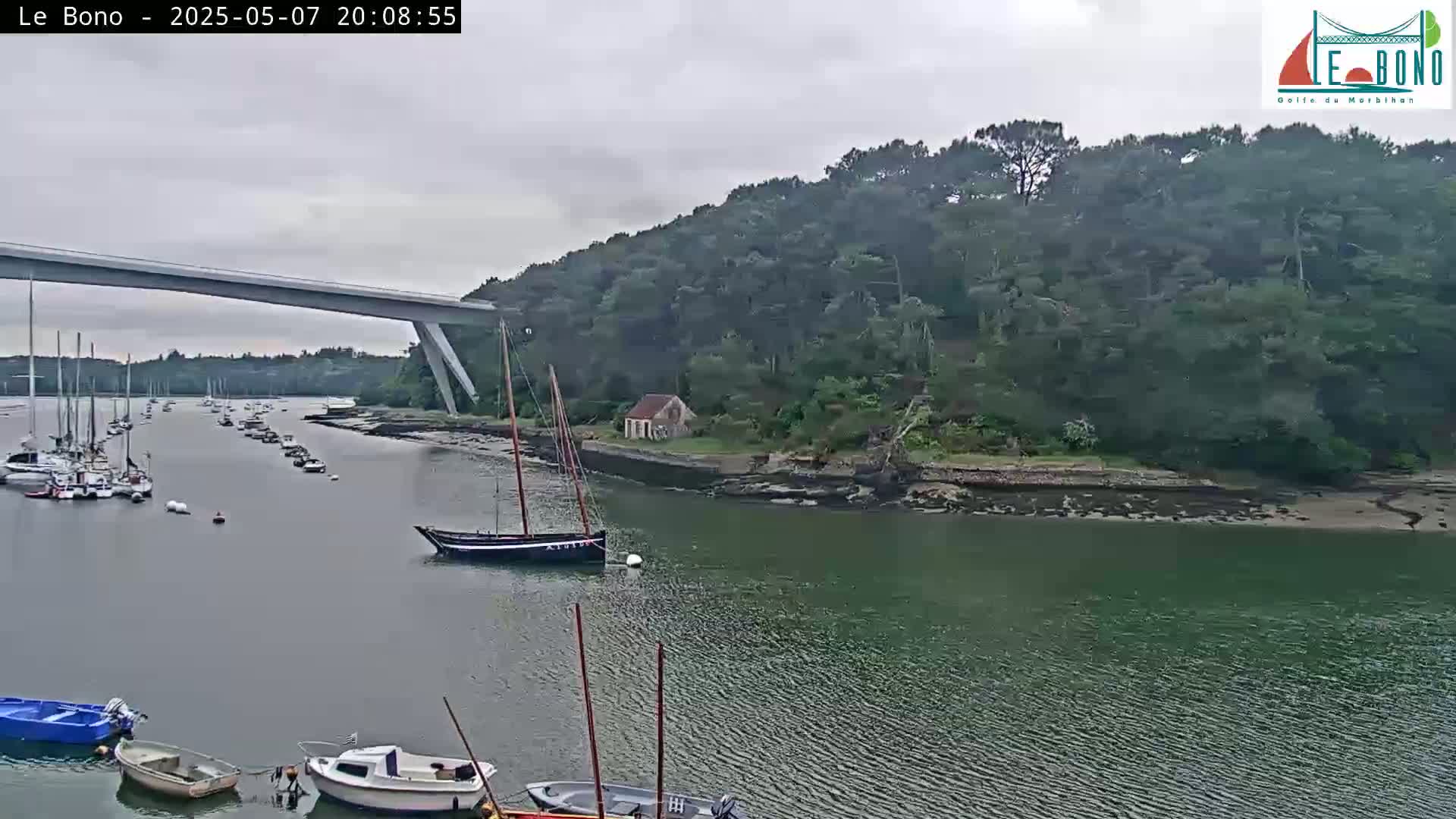 A calm, overcast day shows a waterway with numerous boats moored along its banks, a small cottage nestled on a wooded hillside, and a long bridge spanning the water in the background.