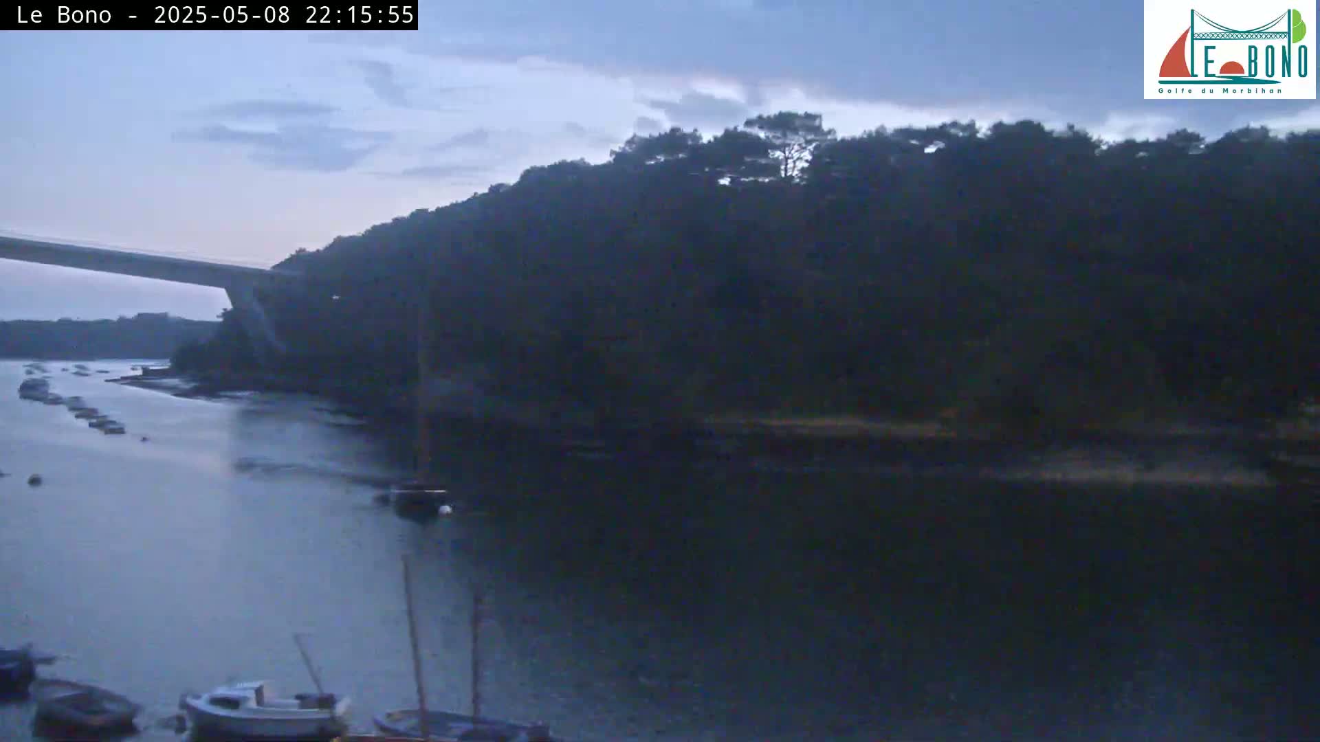 Under a twilight sky, a calm body of water shows several boats moored near a wooded shoreline and a bridge extending across the water.