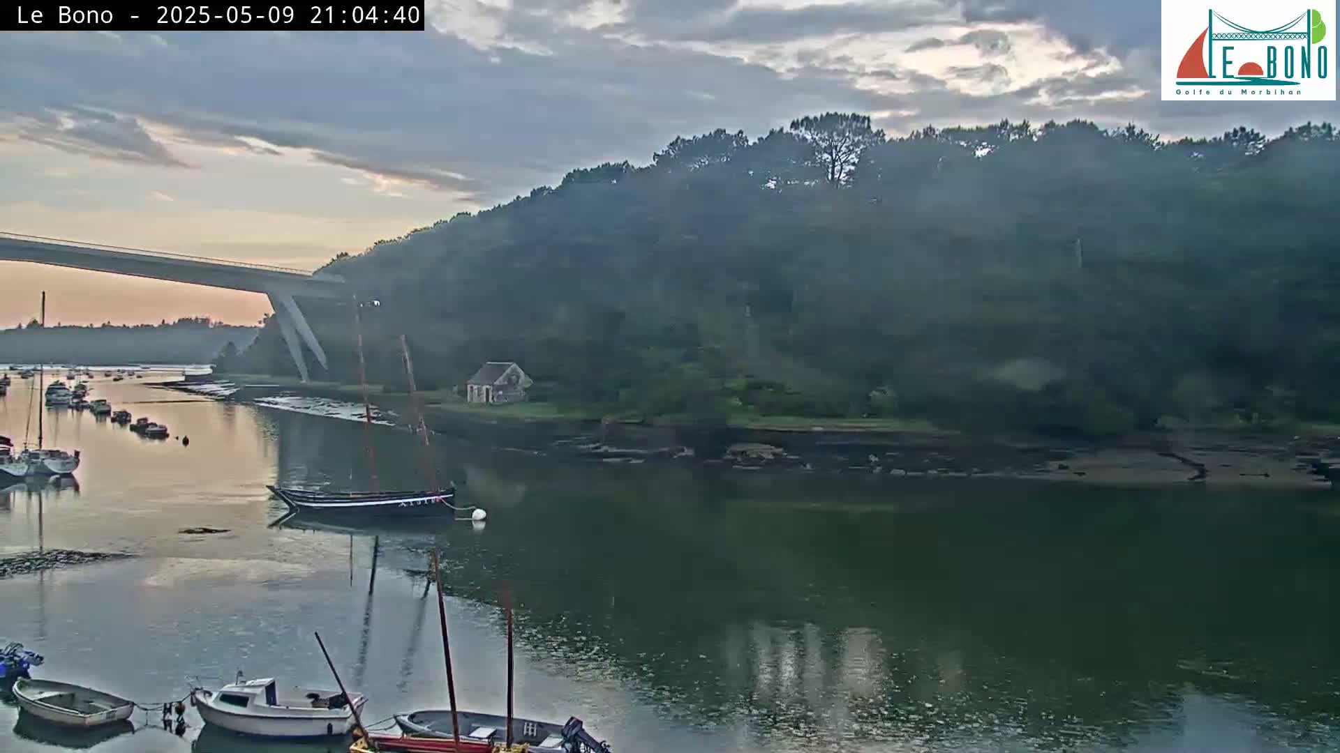 Several boats are moored in a calm river at dusk under an overcast sky, with a bridge and wooded hillside in the background.
