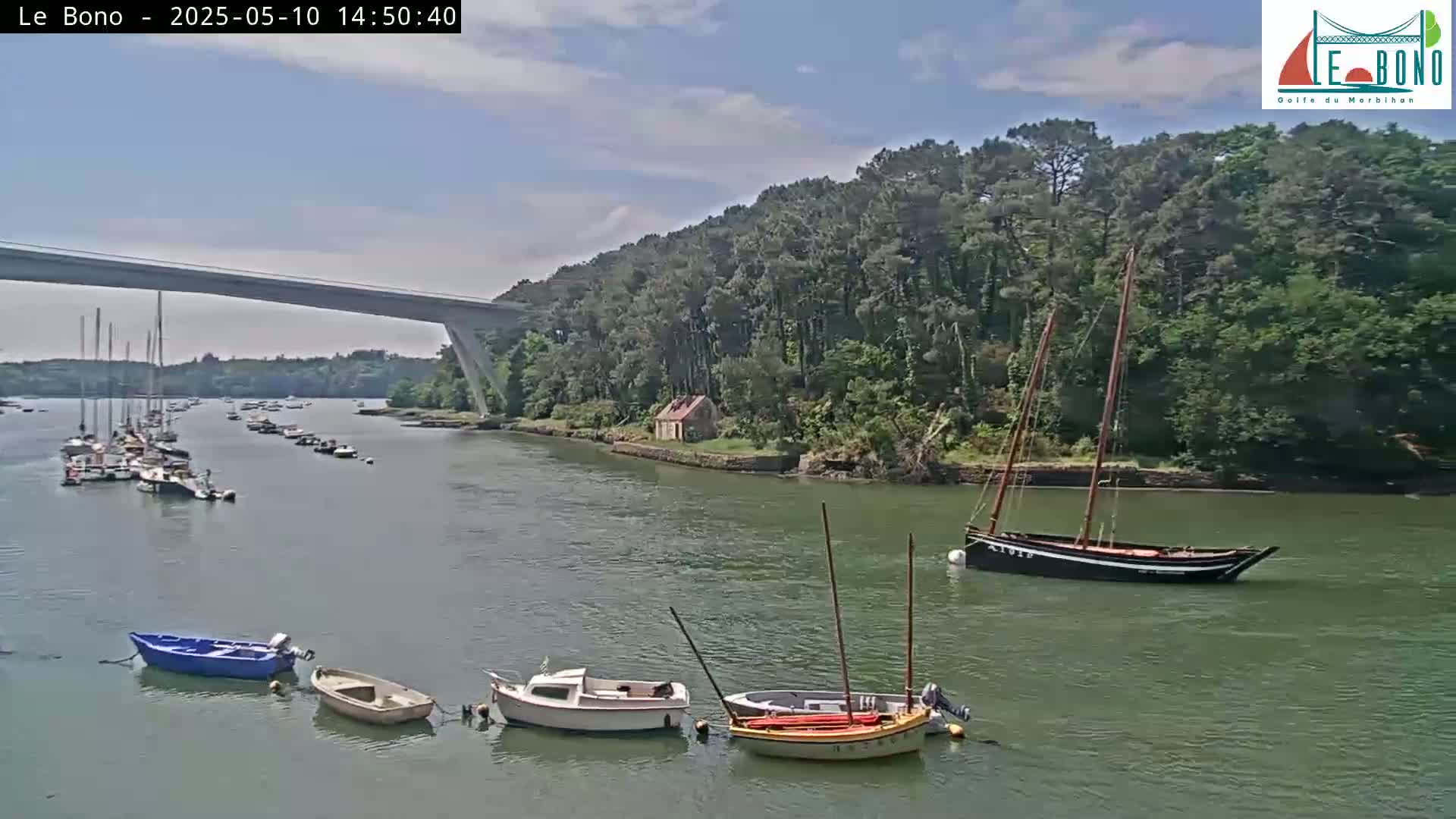 Several small motorboats and sailboats are moored in a calm waterway under a partly cloudy sky, with a bridge spanning the water in the background and a wooded shoreline.