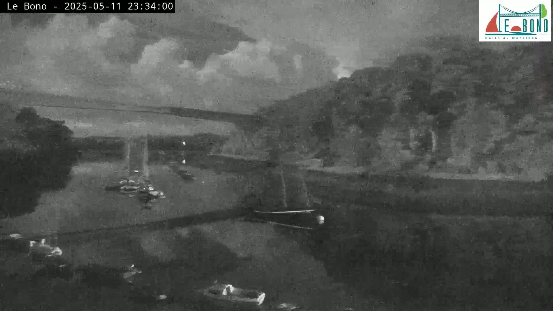 A nighttime, overcast scene shows a body of water with several small boats, a larger boat, and a bridge in the background.