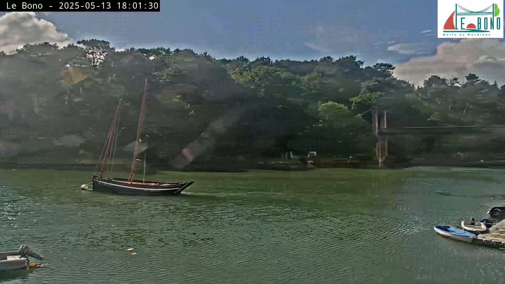 A sailboat and several smaller boats are on calm, dark green water, backed by a lush green hillside under a mostly sunny sky.