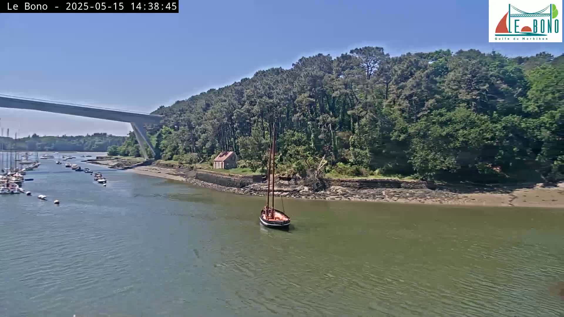 A small sailboat is moored in a calm waterway under a sunny sky, near a bridge and a wooded shoreline with several other boats in the distance.