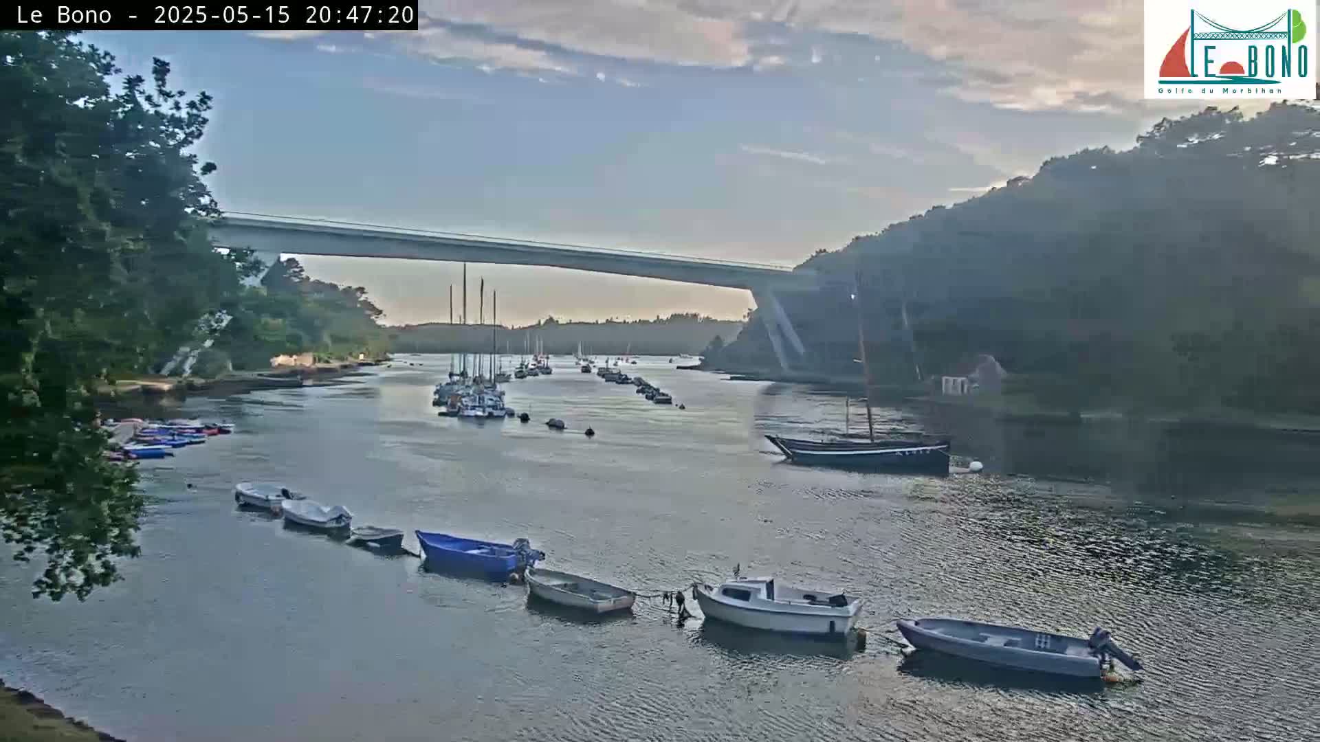 Under a partly cloudy sky, a river scene shows numerous small boats moored along the shore and others anchored further out, with a long bridge spanning the waterway in the background.