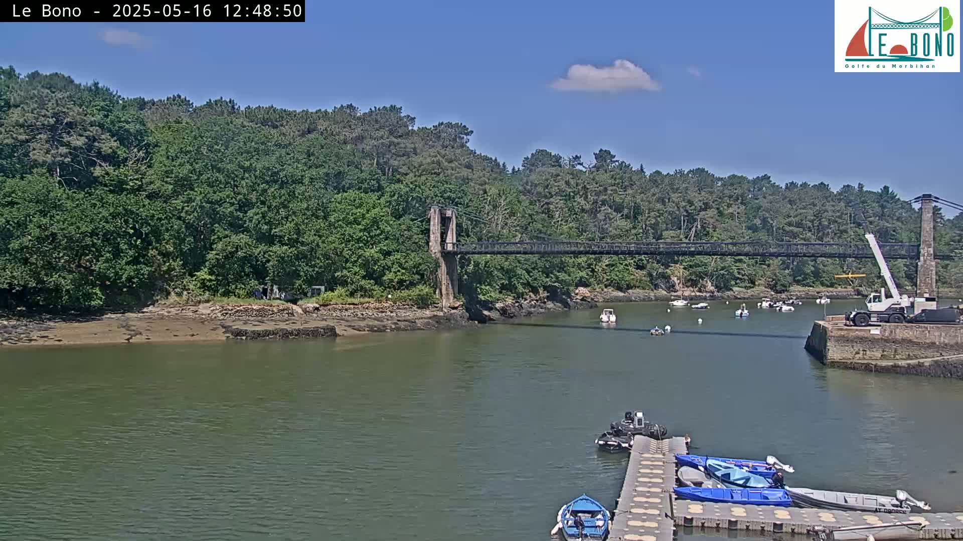 A suspension bridge spans a calm river under a mostly sunny sky, with several small boats moored on a dock and others in the water.
