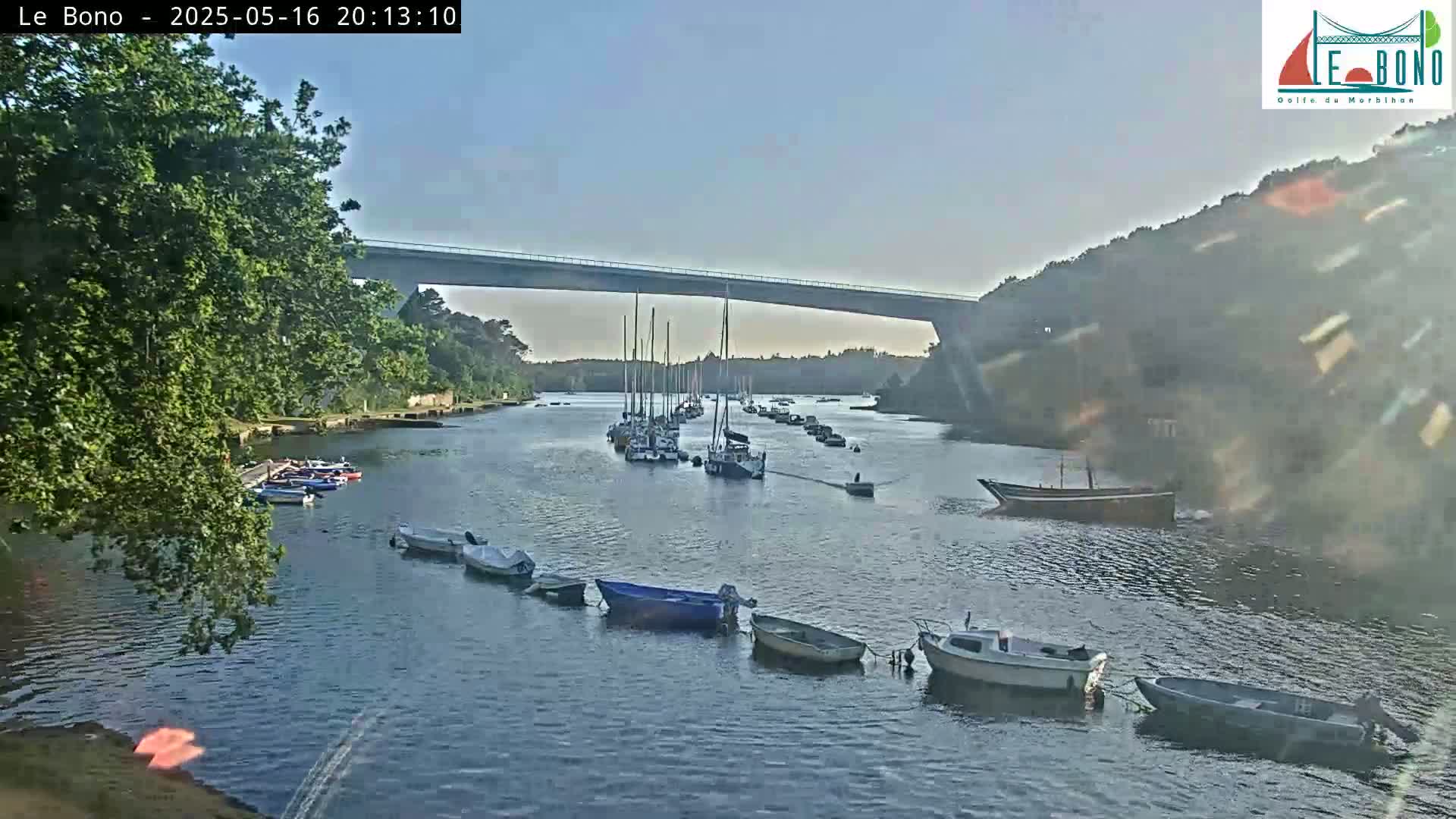Under a clear sky, numerous boats are moored along a calm waterway that passes under a long bridge, with lush green trees lining the banks.