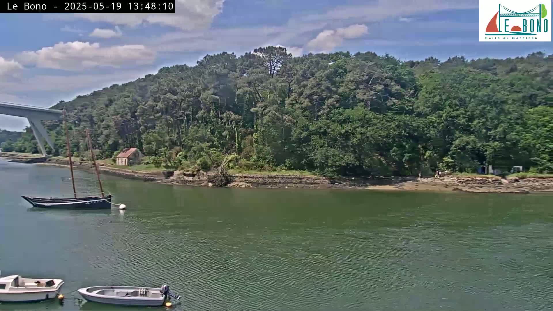 A partly cloudy sky overlooks a calm body of water where several small boats are moored near a wooded shoreline with a small house.