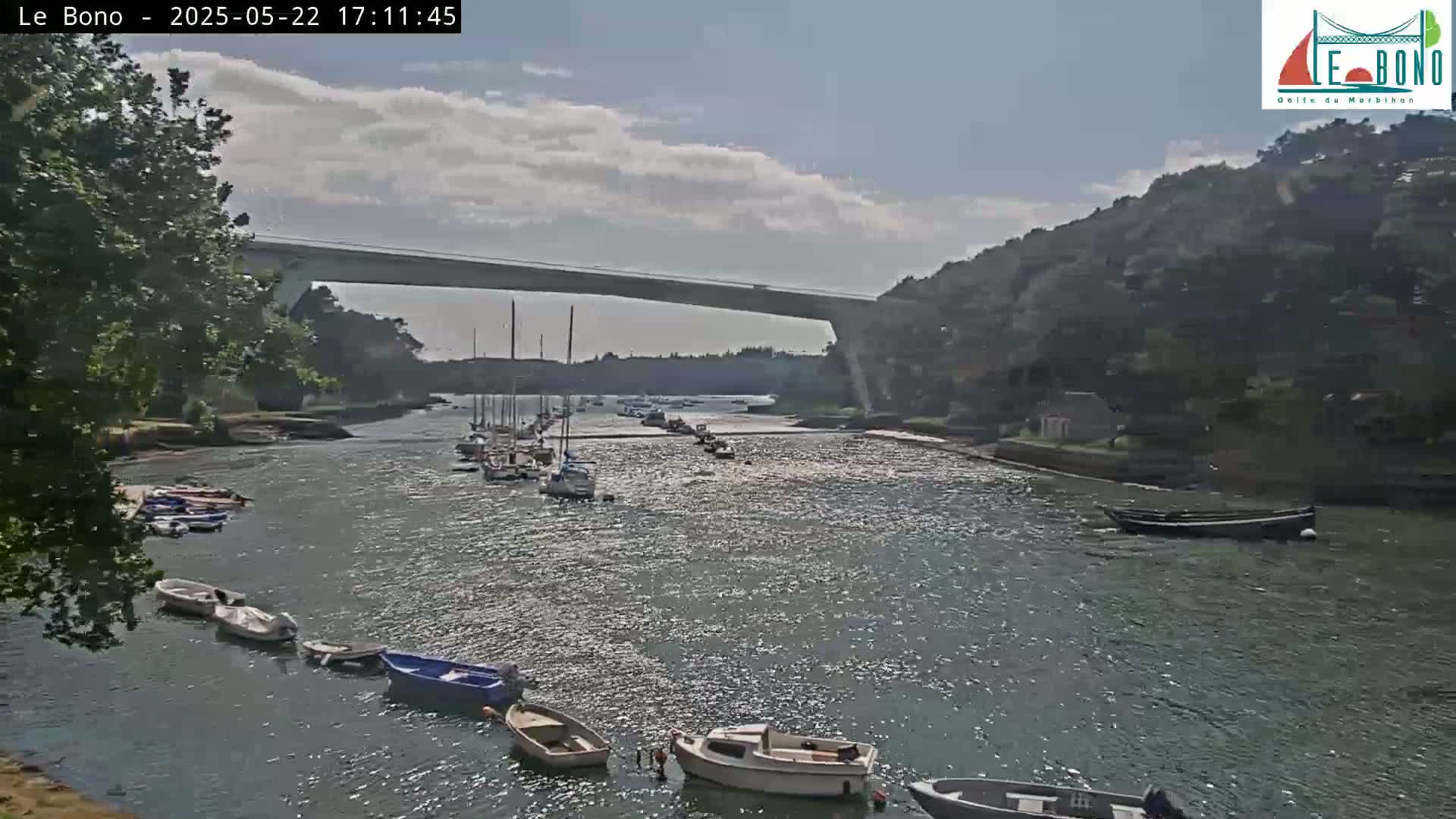 A partly cloudy day shows a waterway with numerous small boats, some moored and others underway, passing under a long bridge flanked by green hills.