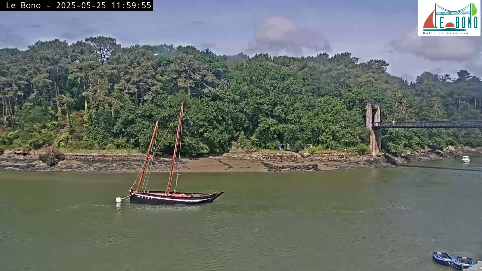A sailboat is moored in calm water near a wooded shoreline and a suspension bridge under a partly cloudy sky.