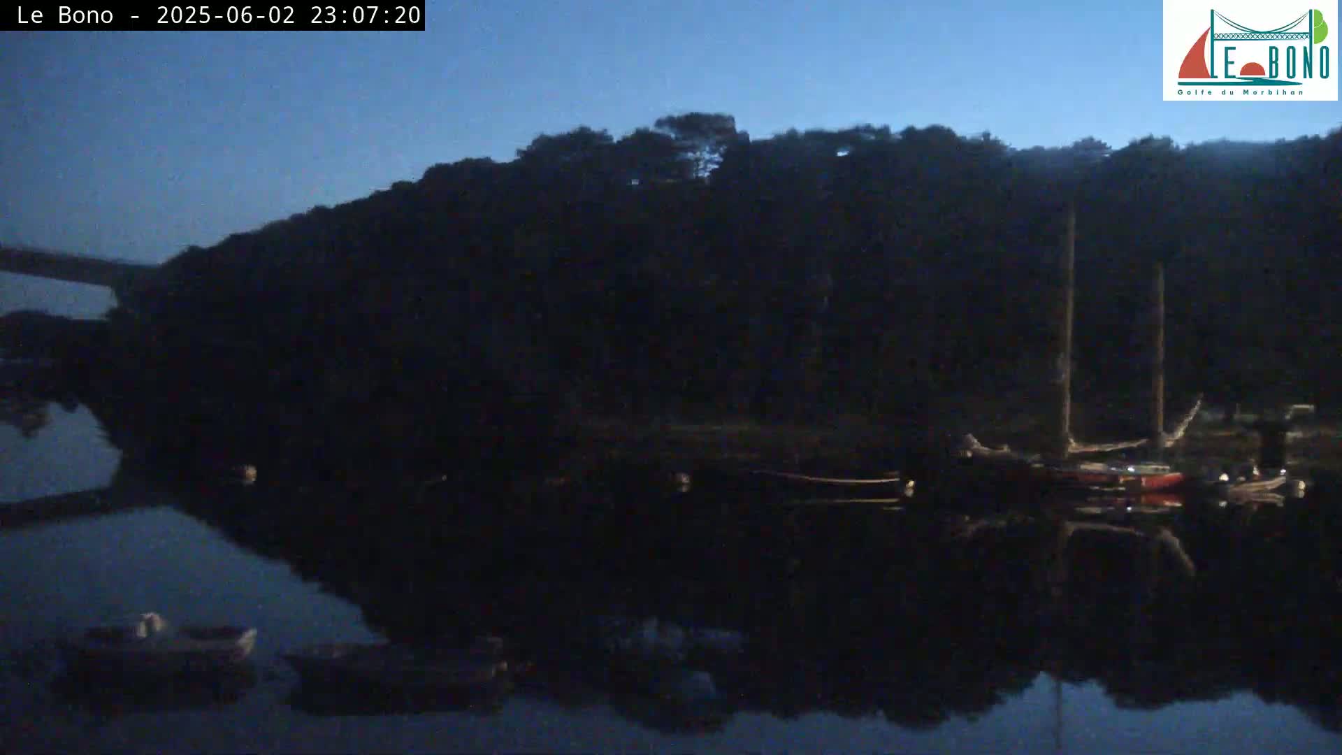 Under a dark blue night sky, several boats are moored along a dark shoreline, reflected in the still water of a body of water.