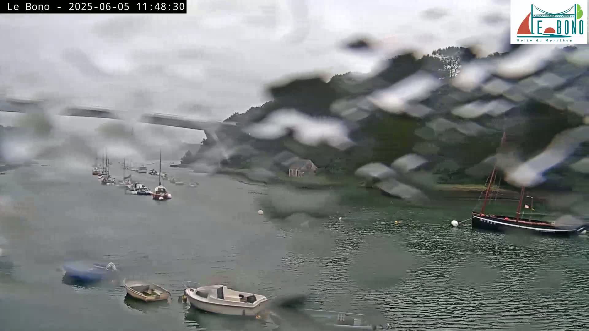 Several boats are moored in a calm waterway under a rainy, overcast sky, viewed through rain-covered glass.