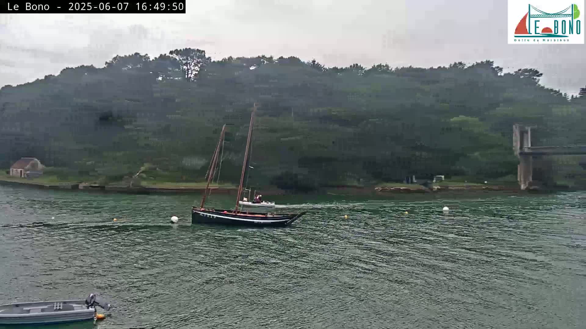 A sailboat is sailing on a calm, overcast body of water near a lush green hill and a bridge.