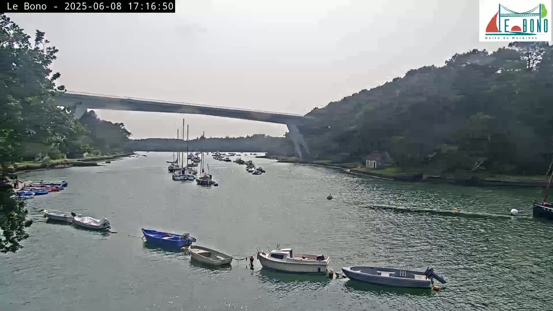 A calm, overcast day shows a waterway with numerous small boats and sailboats moored near a high bridge spanning a narrow channel between wooded banks.