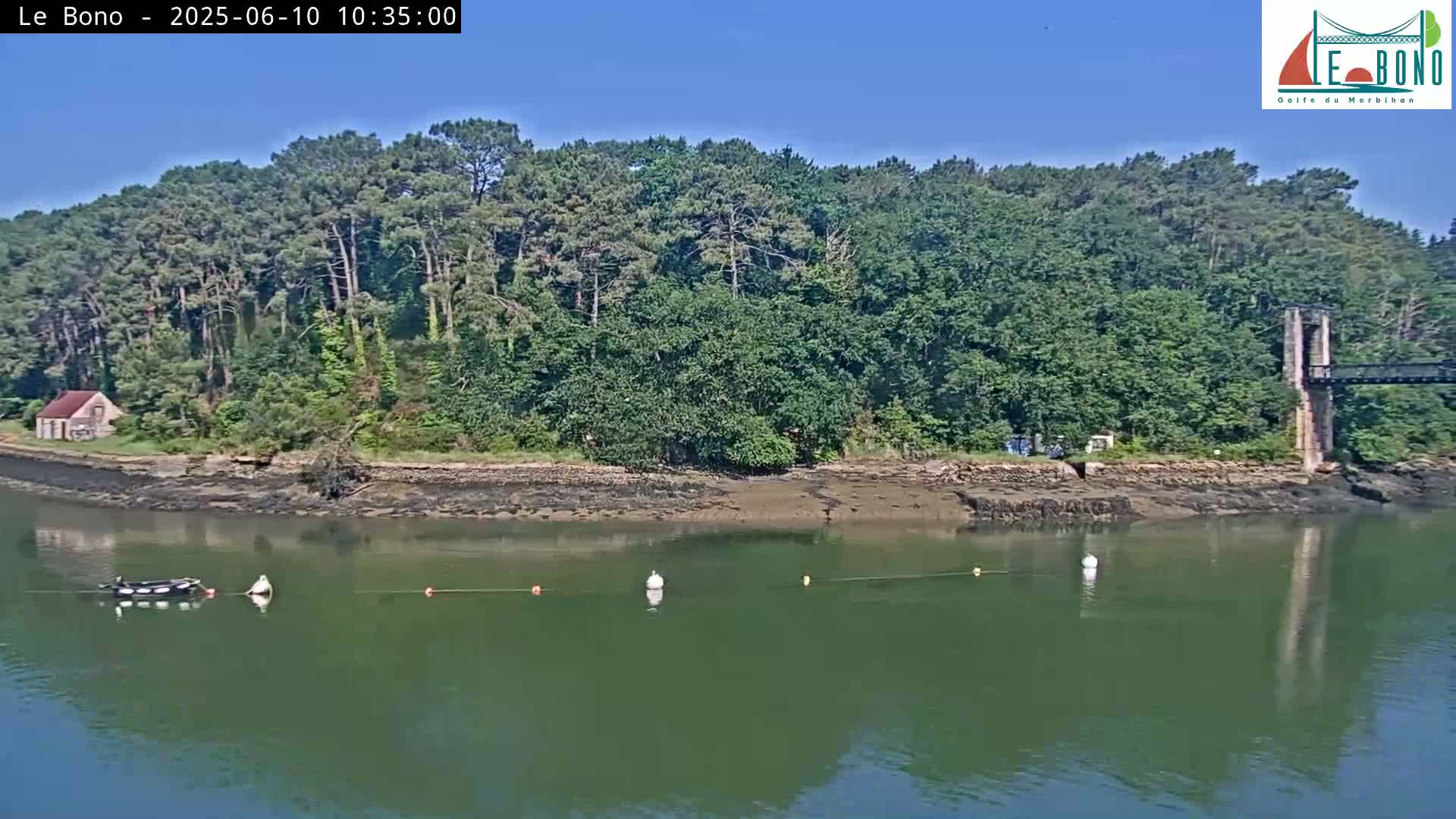 A small boat is moored in calm, green water next to a shoreline with lush green trees, a small building, and part of a stone bridge visible in the background under a clear blue sky.
