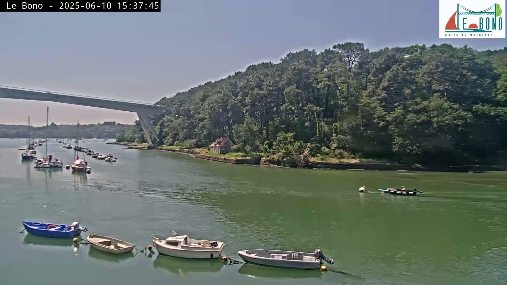 A calm, sunny day shows a waterway with numerous small boats, a bridge in the distance, and a wooded shoreline.