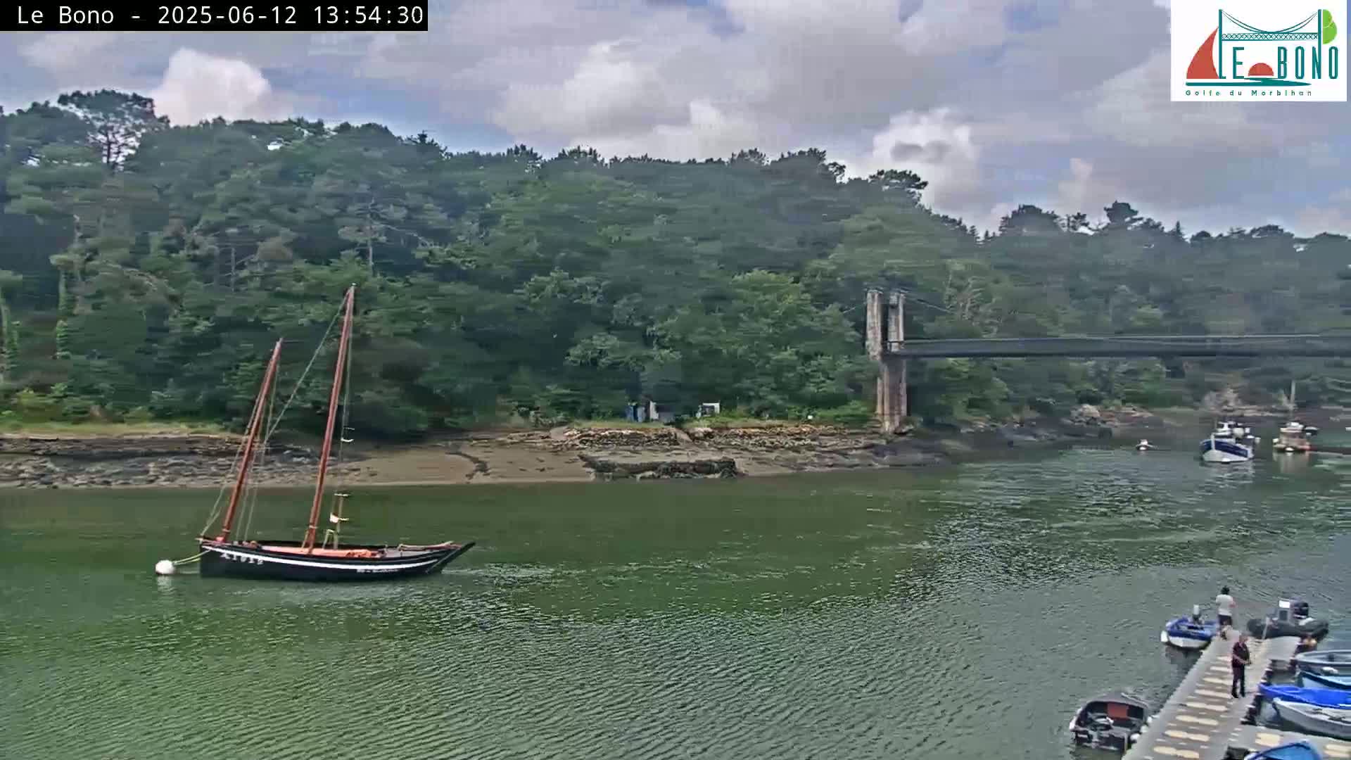 A sailboat is moored in a calm waterway under a cloudy sky, near a suspension bridge and a shoreline lined with green trees.