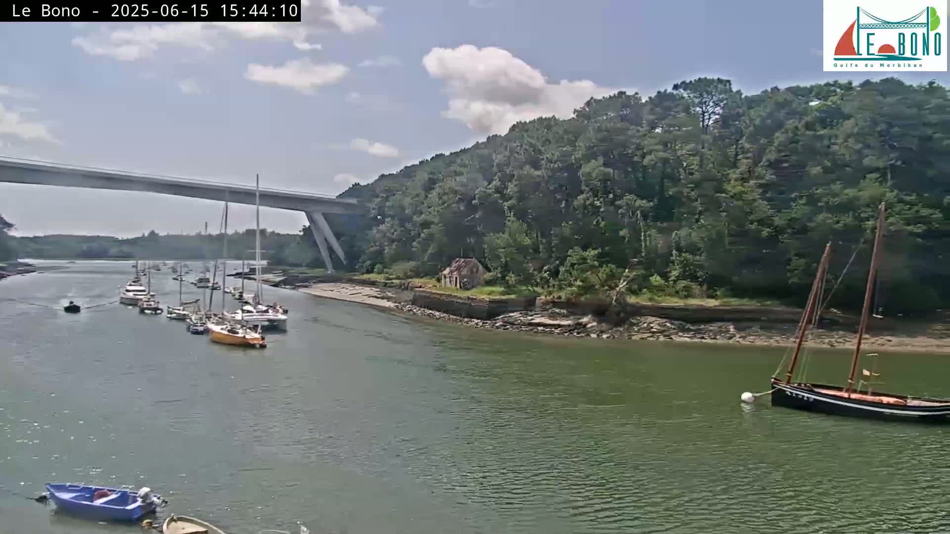 Under a partly cloudy sky, several sailboats are moored in a calm waterway under a long bridge, with a small dilapidated building visible on the shoreline.